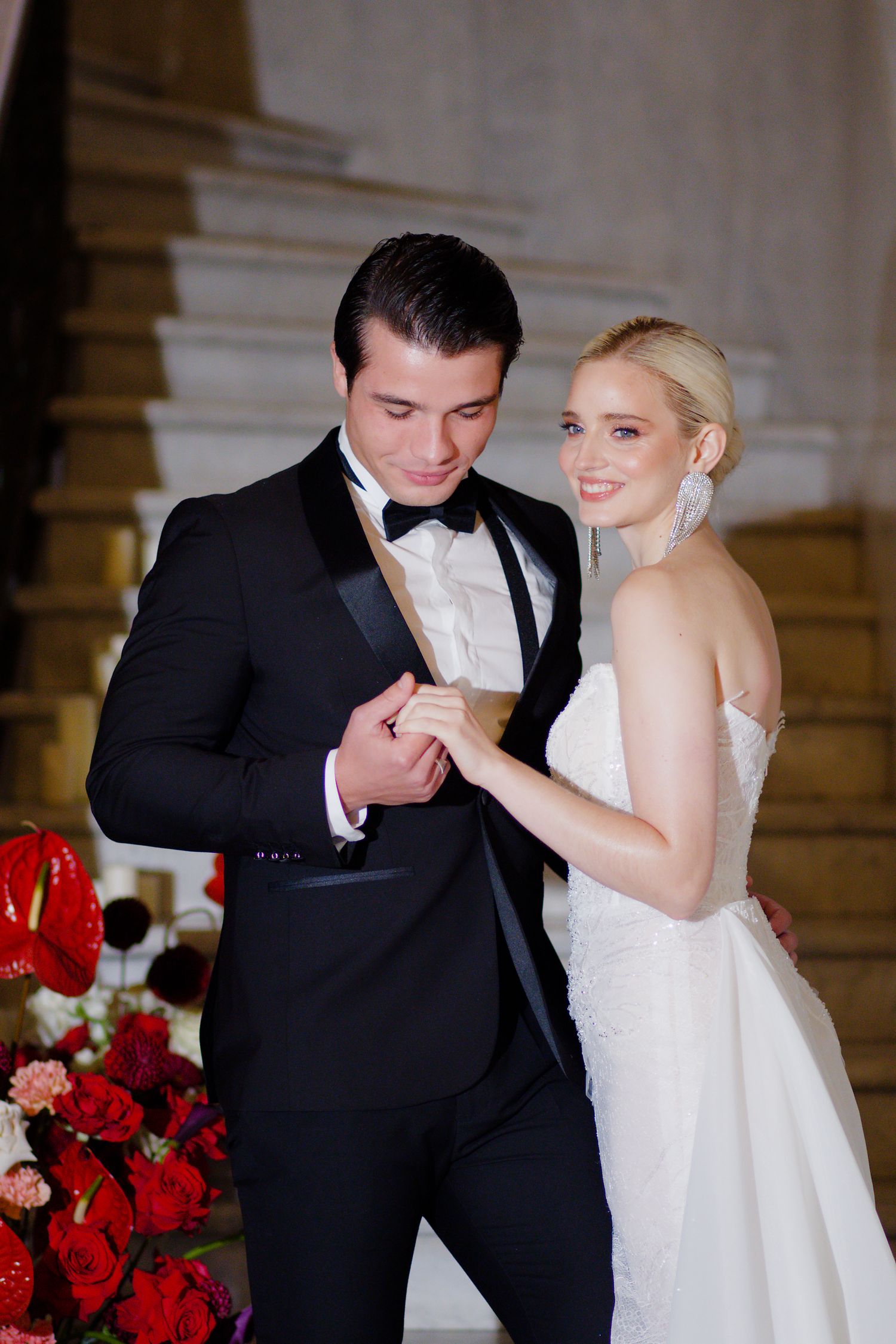 A wedding couple shares an intimate moment near red rose arrangements on marble steps.