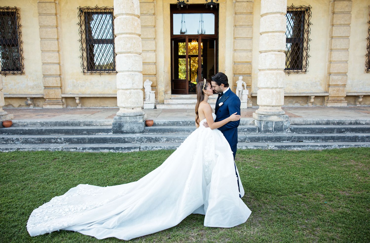 A bride and groom embrace on a manicured lawn in front of a grand historic building.