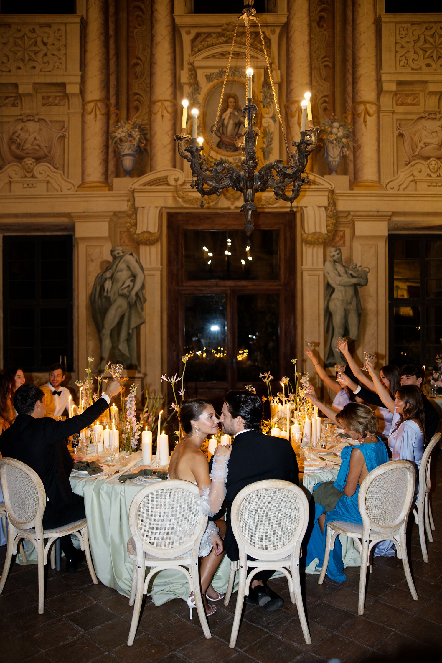 Wedding guests celebrate at an ornate venue with grand chandeliers, statues, and candlelit tables during an evening reception.