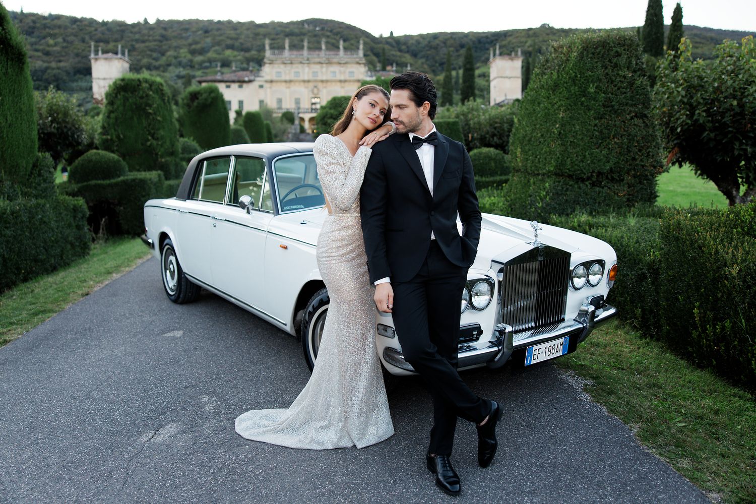 A couple poses by a classic white Rolls-Royce against a backdrop of an elegant mansion and manicured gardens.