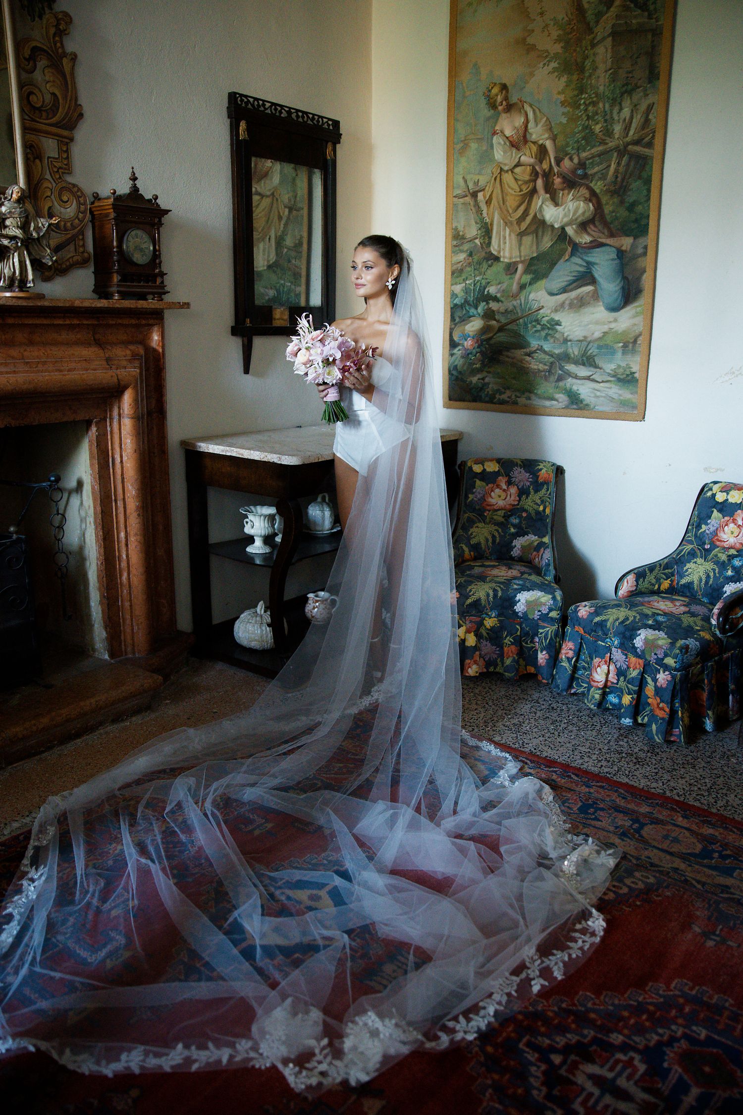 A bride in a flowing cathedral veil poses elegantly in a vintage room with ornate decor and classical artwork.