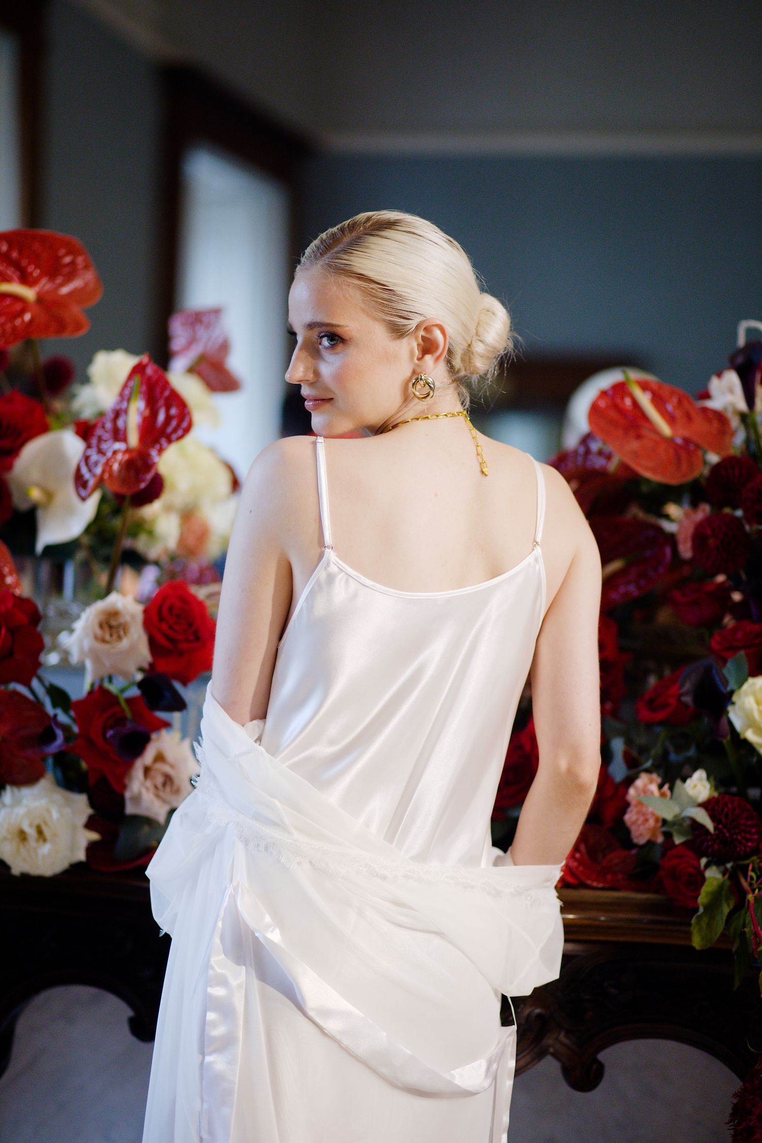 Elegant bride in white dress stands amid vibrant red and white floral arrangements in a dim-lit room.