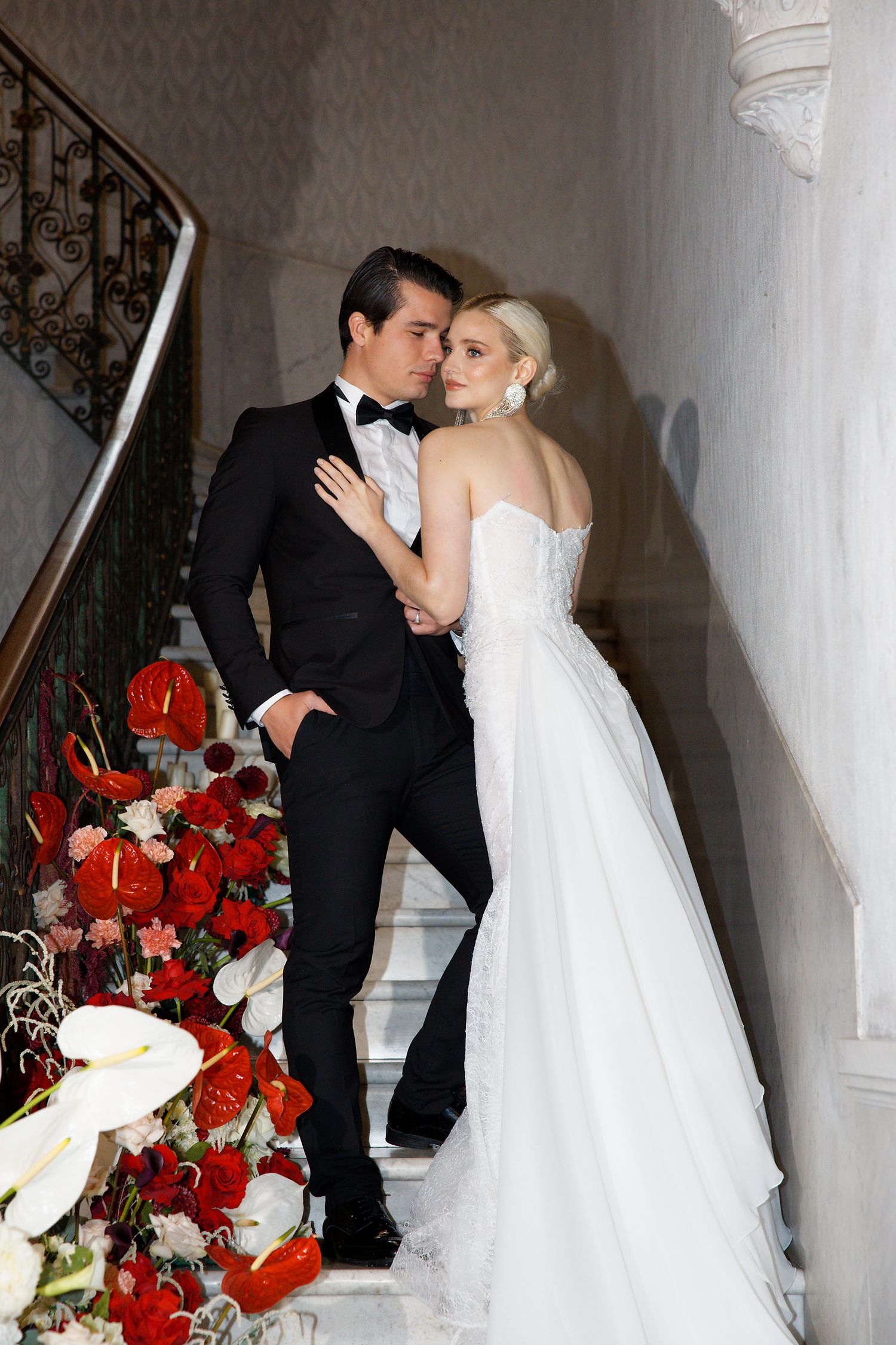 Elegant couple posing on staircase with red floral arrangements.