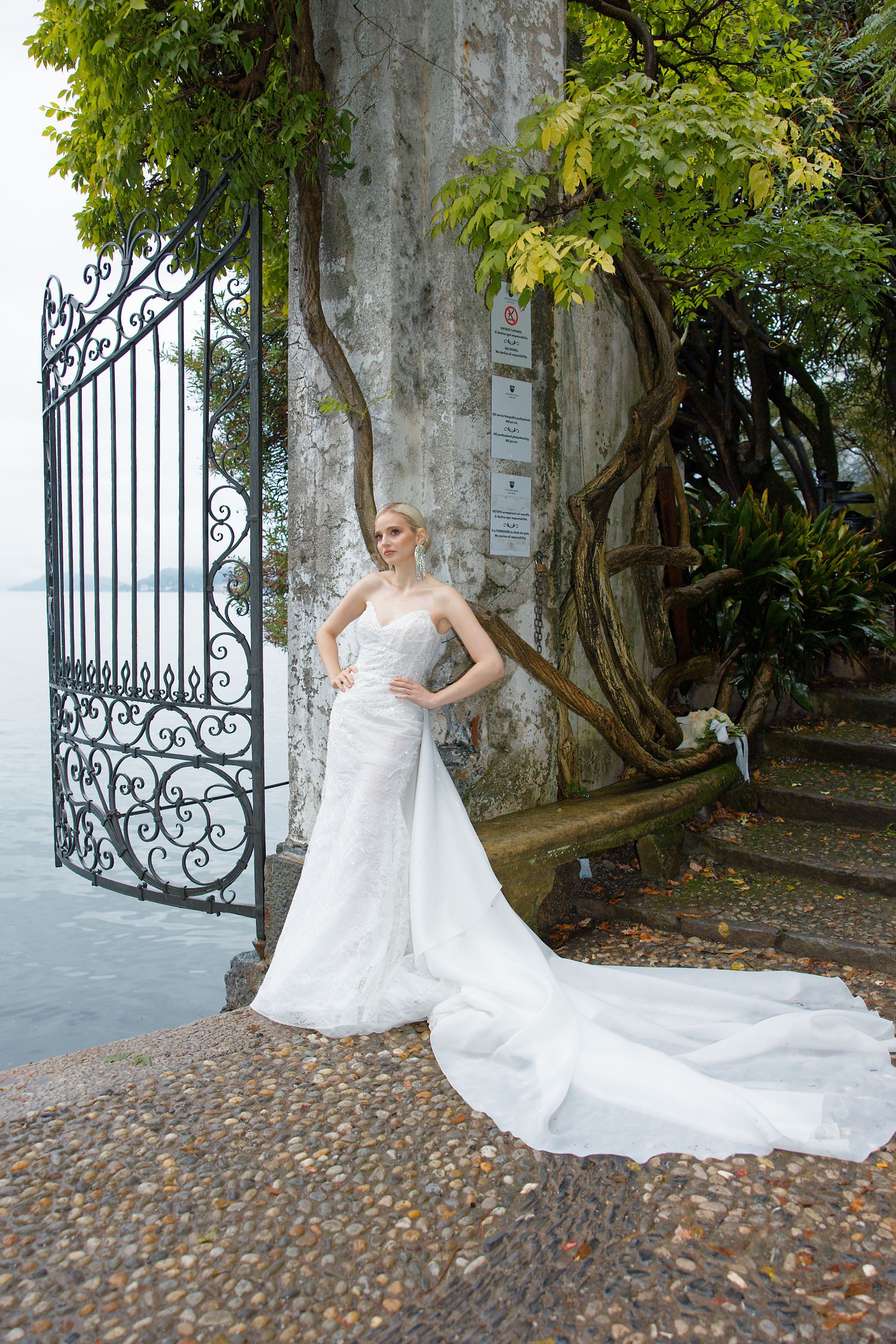 A bride in a flowing white gown poses elegantly by an ornate iron gate with a scenic backdrop.