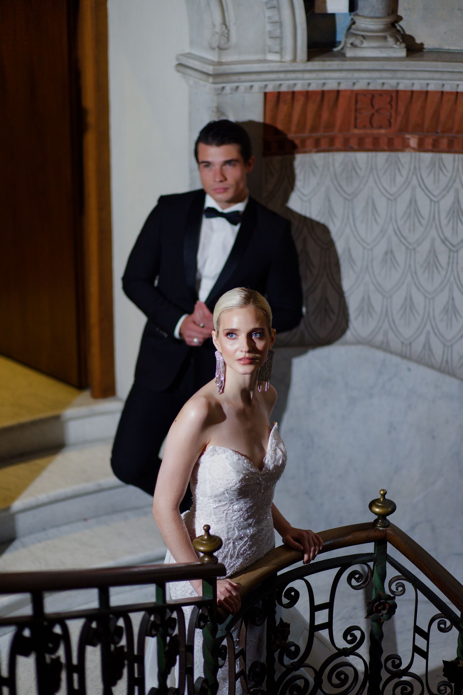 An elegant wedding couple poses on an ornate marble staircase with decorative iron railing.