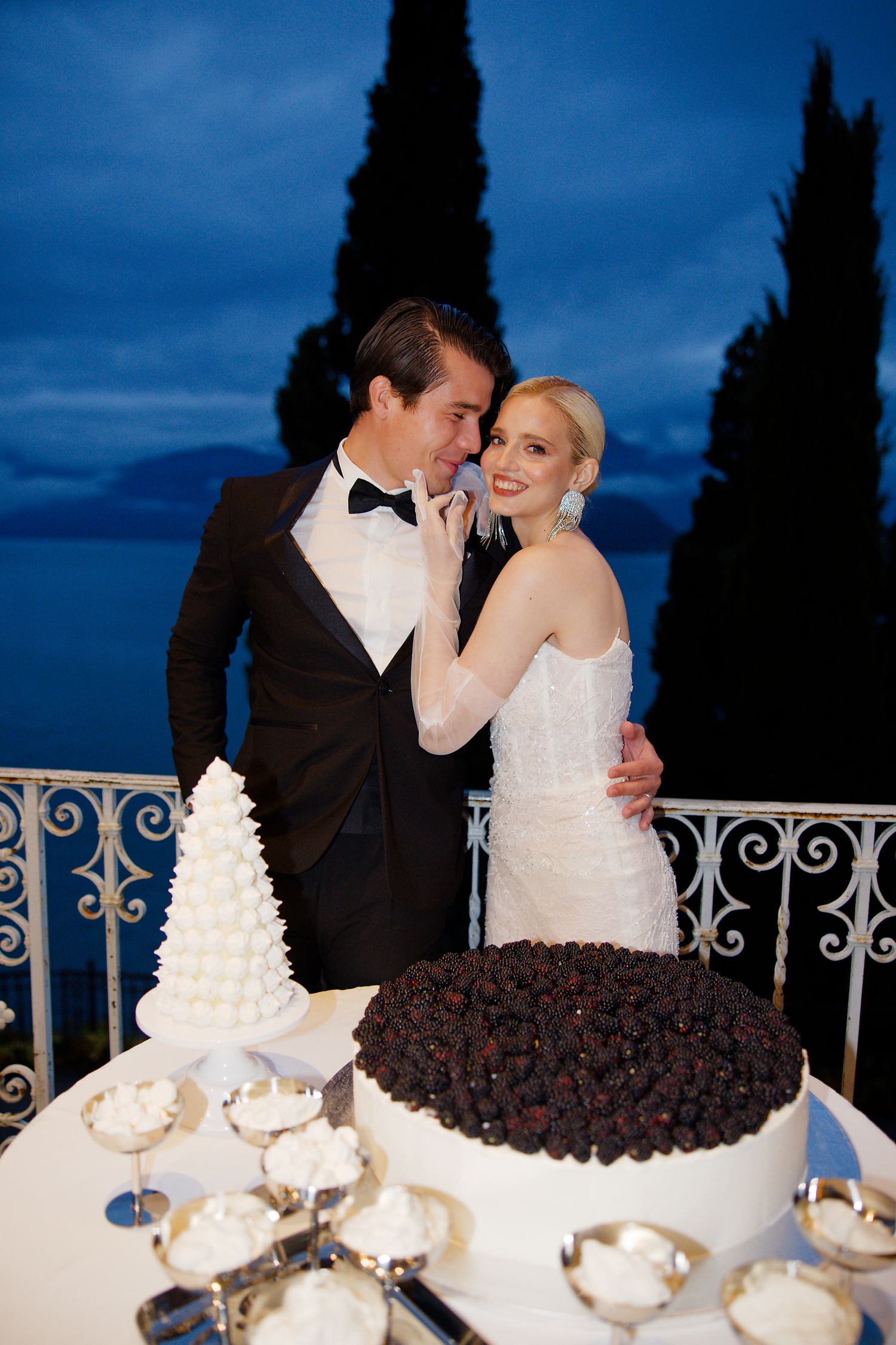 A romantic wedding moment captured at dusk with cypress trees and a two-tiered cake display.