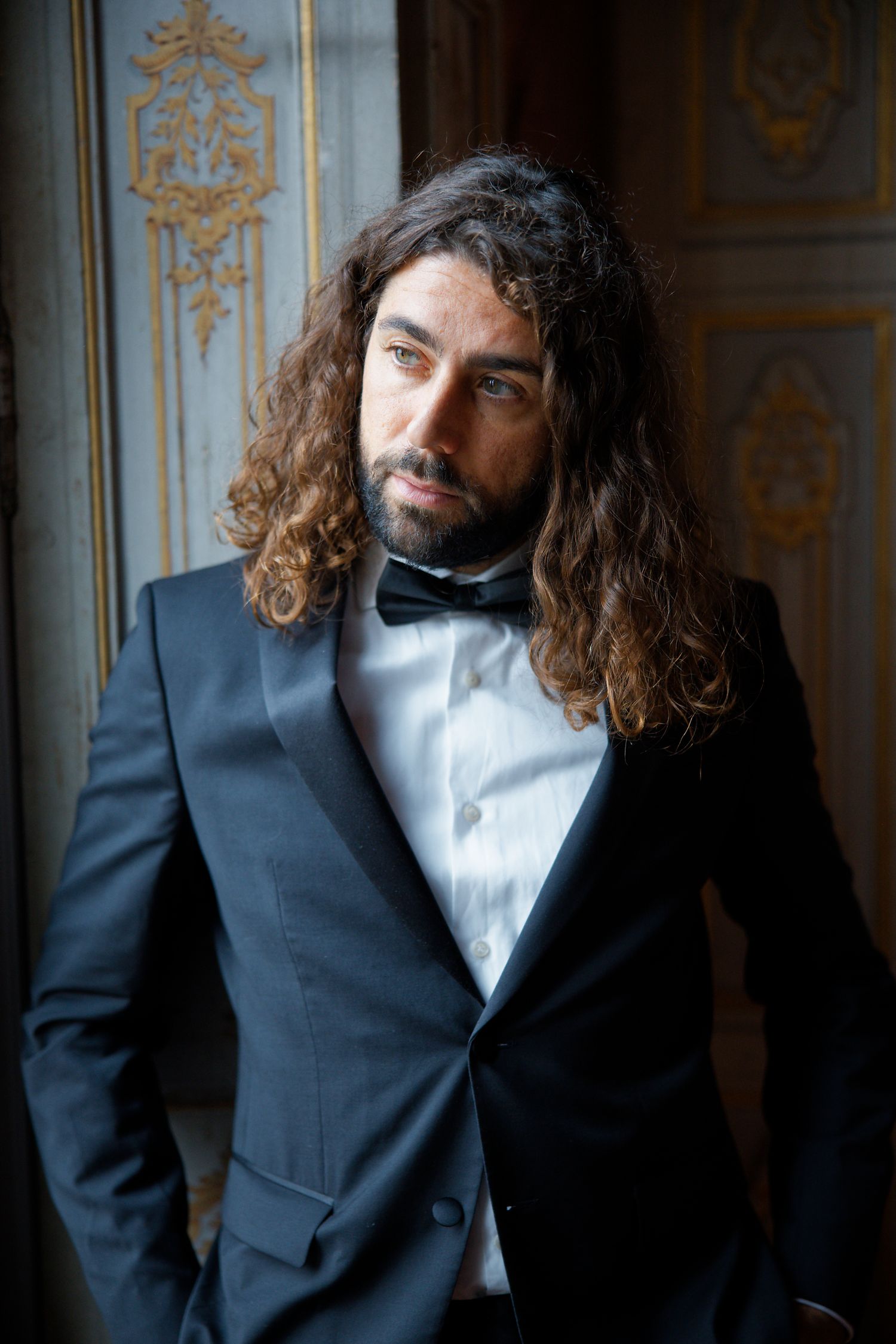 Long-haired groom in black tuxedo and bowtie poses by ornate wall.