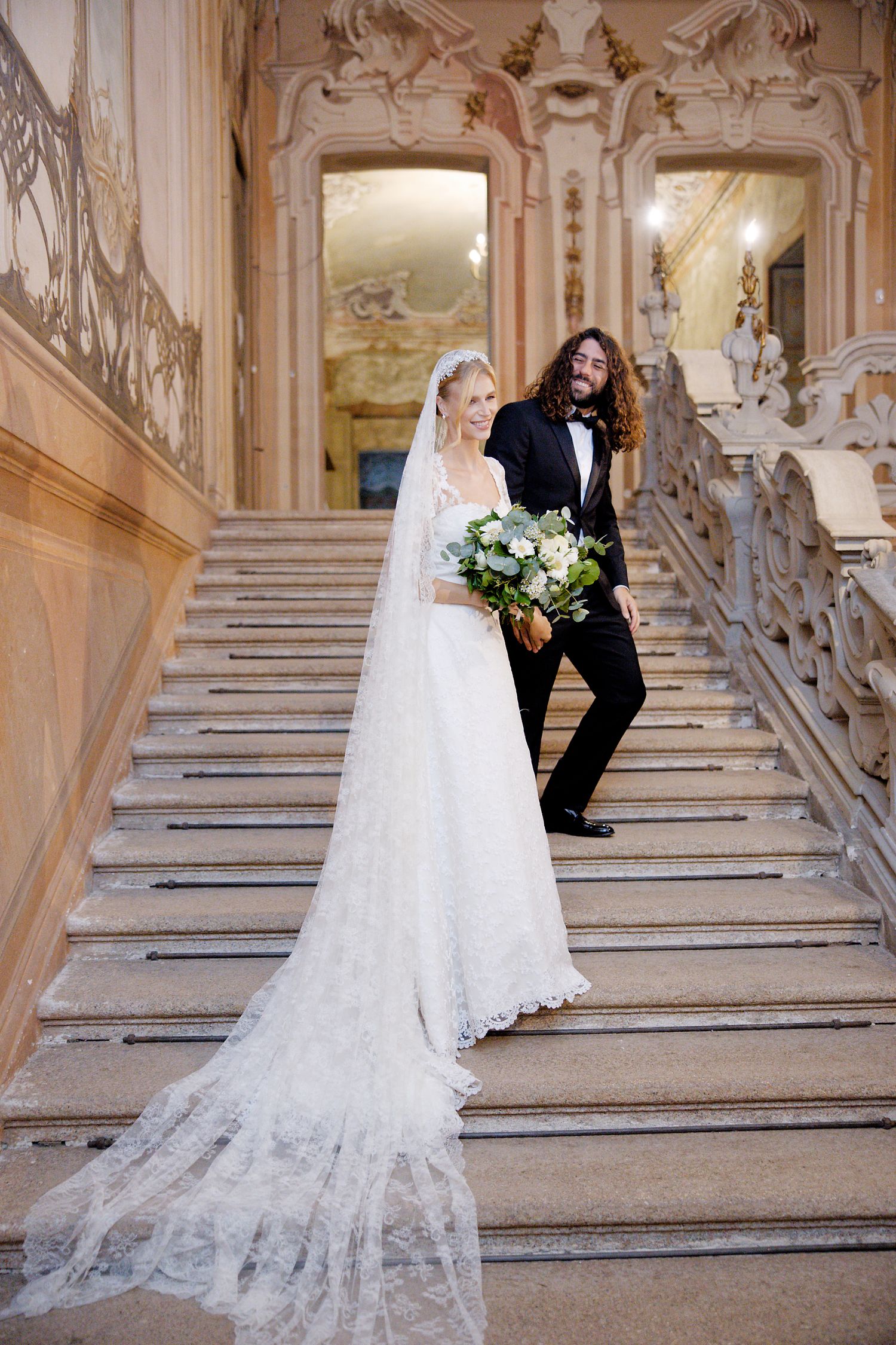 A couple on ornate marble stairs in a historic palace setting, with the bride wearing a flowing white gown and long veil.