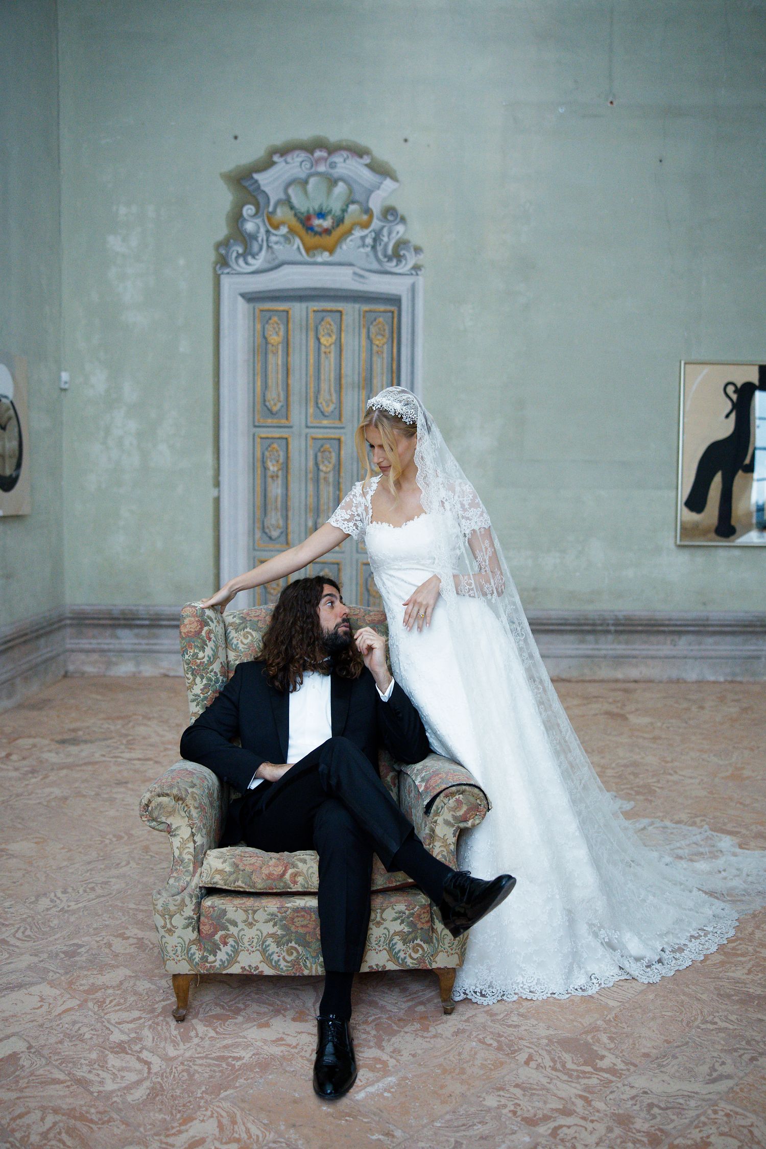 A bride in a white lace gown and veil stands beside someone in a black tuxedo lounging in a vintage floral armchair.