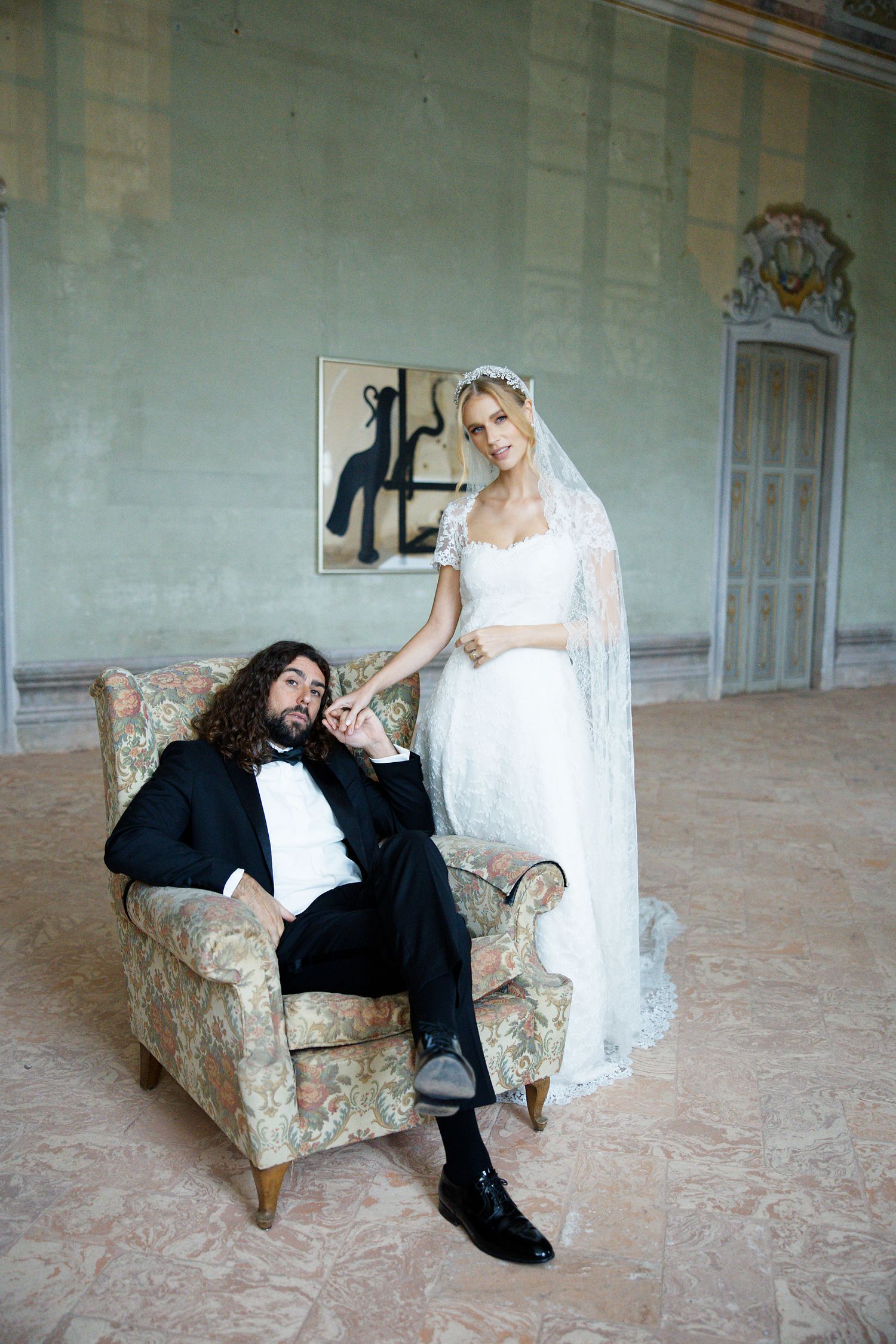A couple in wedding attire poses in an elegant room with a floral armchair and decorative wall art.