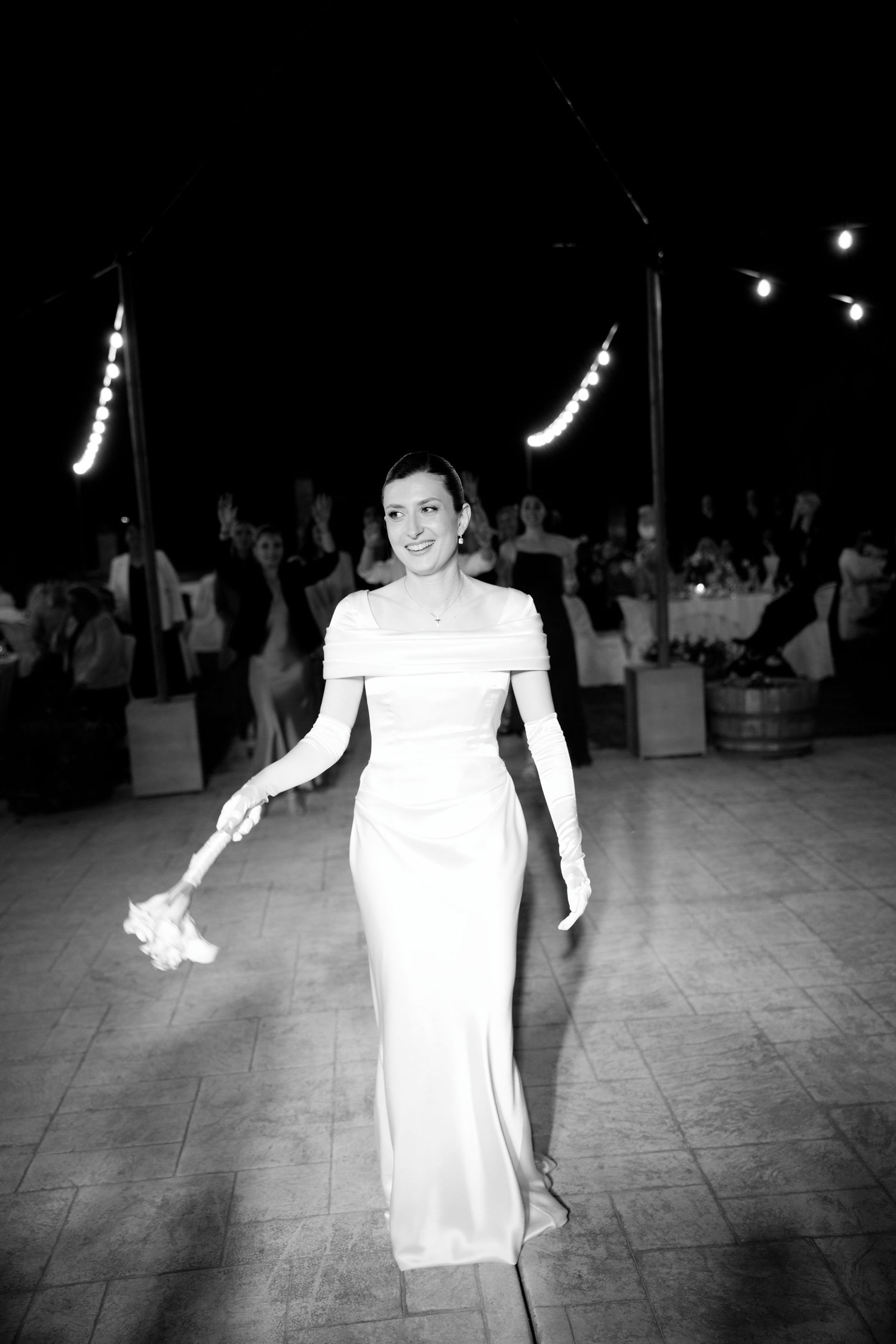 A bride in an elegant white form-fitting gown walks gracefully across a dance floor at a wedding reception.