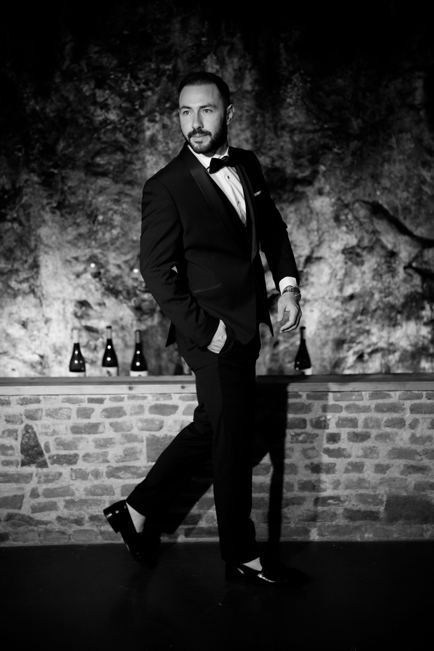 A groom in a tuxedo poses playfully against a rustic brick wall backdrop.