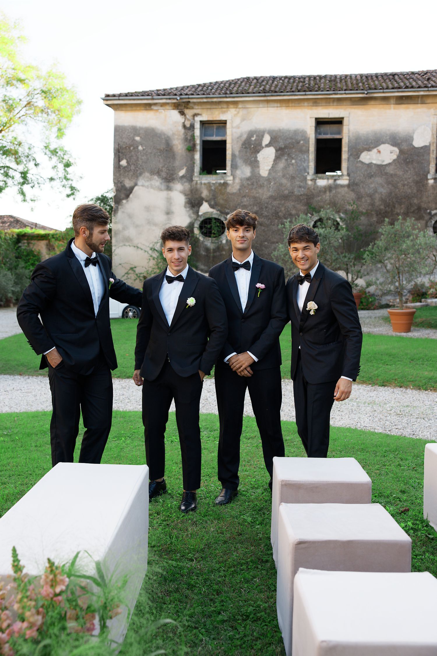 Groomsmen in black tuxedos stand together on a lawn outside a rustic Italian villa.