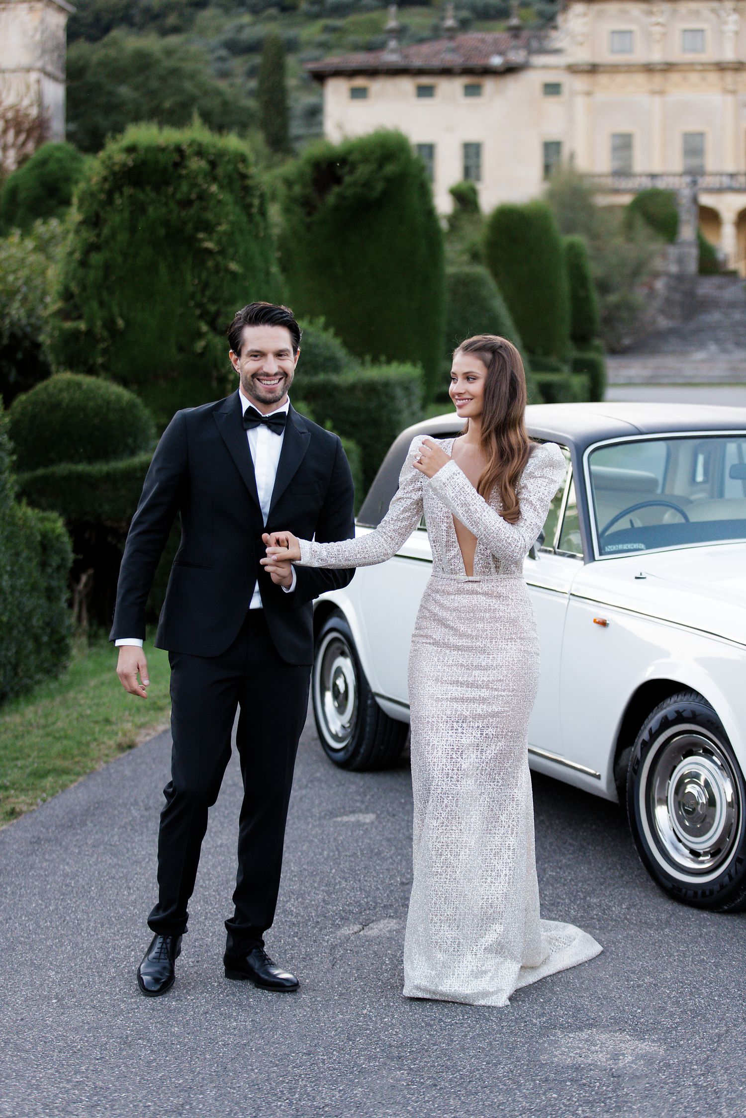 A couple in wedding attire poses elegantly next to a classic white car with manicured topiary gardens in the background.