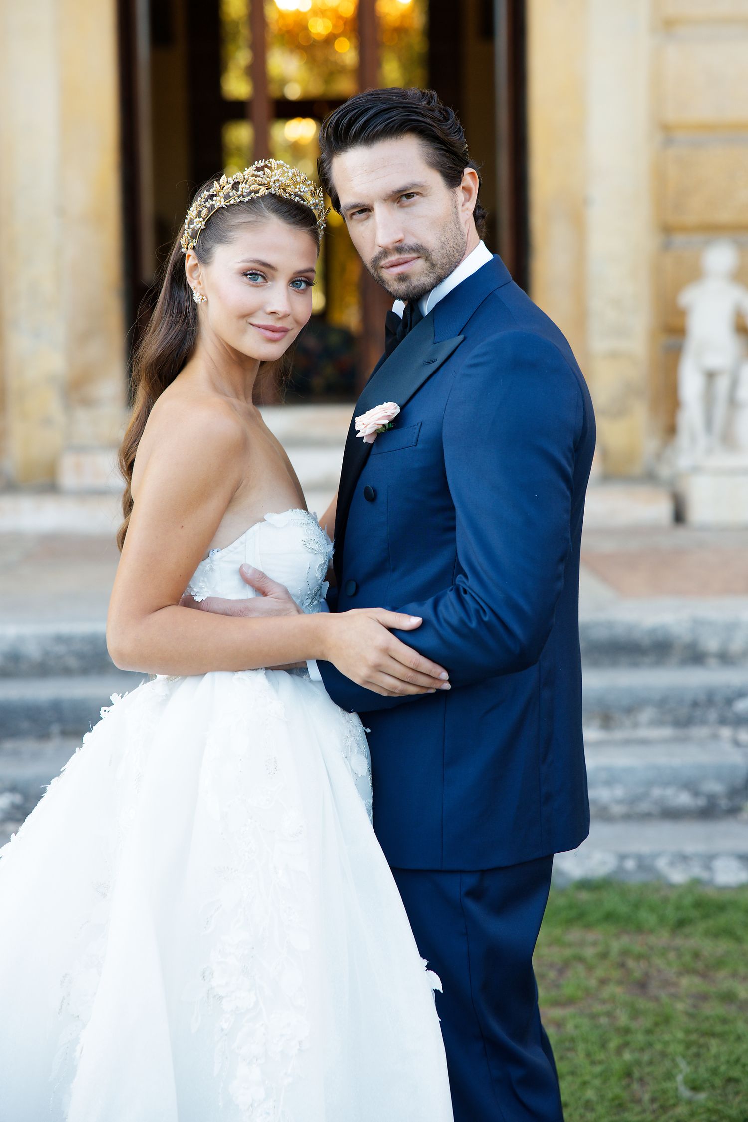 An elegant wedding portrait featuring a bride in white gown and groom in navy suit outside a historic building.
