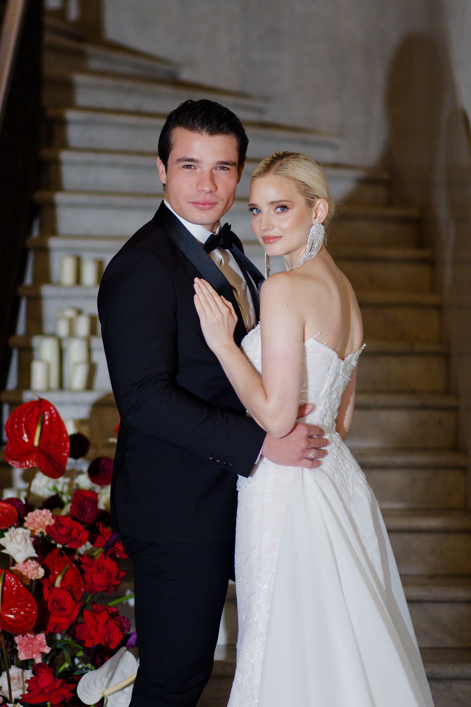 Wedding couple poses formally on elegant staircase with red rose decorations.
