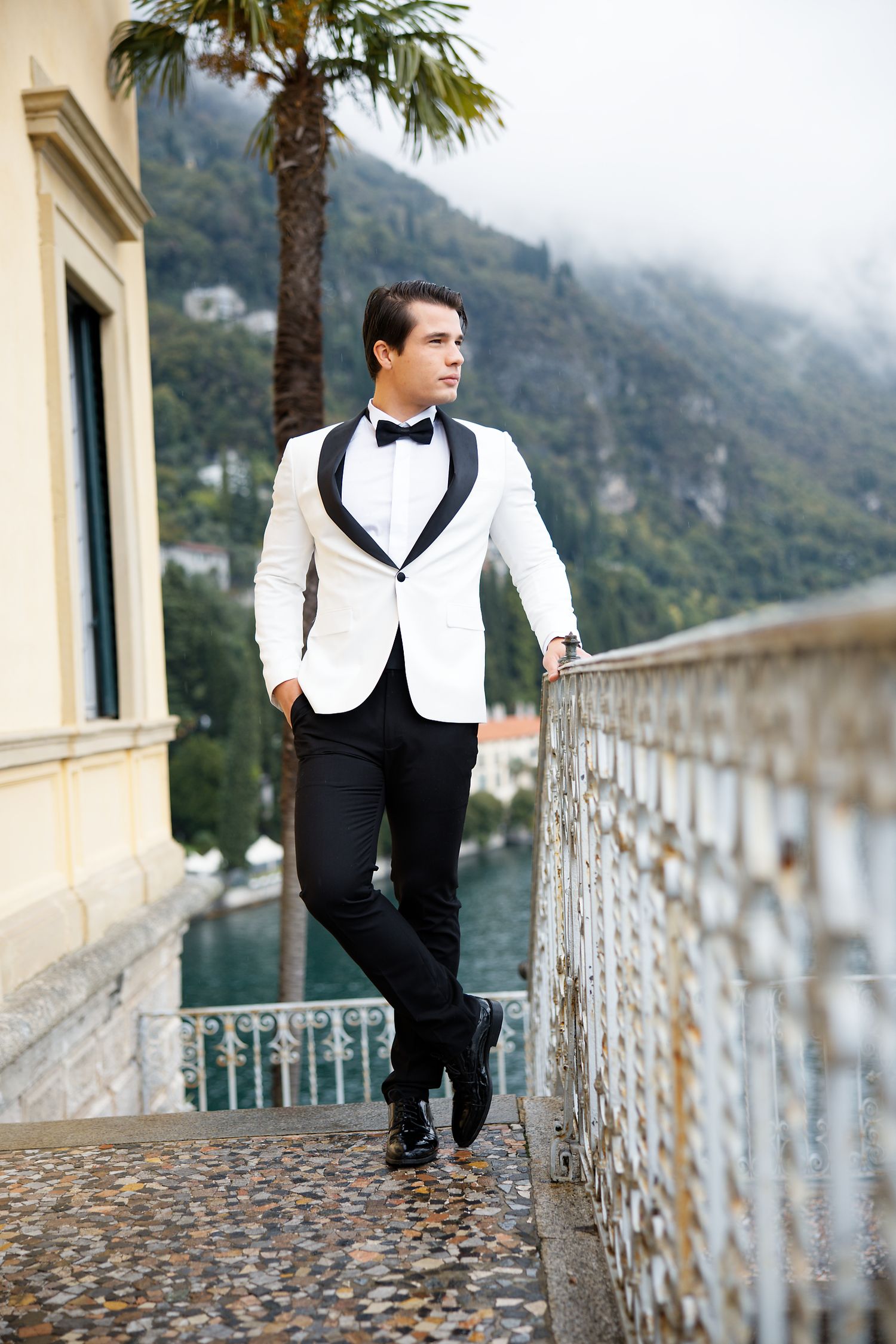 Stylish groom in white tuxedo jacket poses on Mediterranean balcony with mountain and sea views.