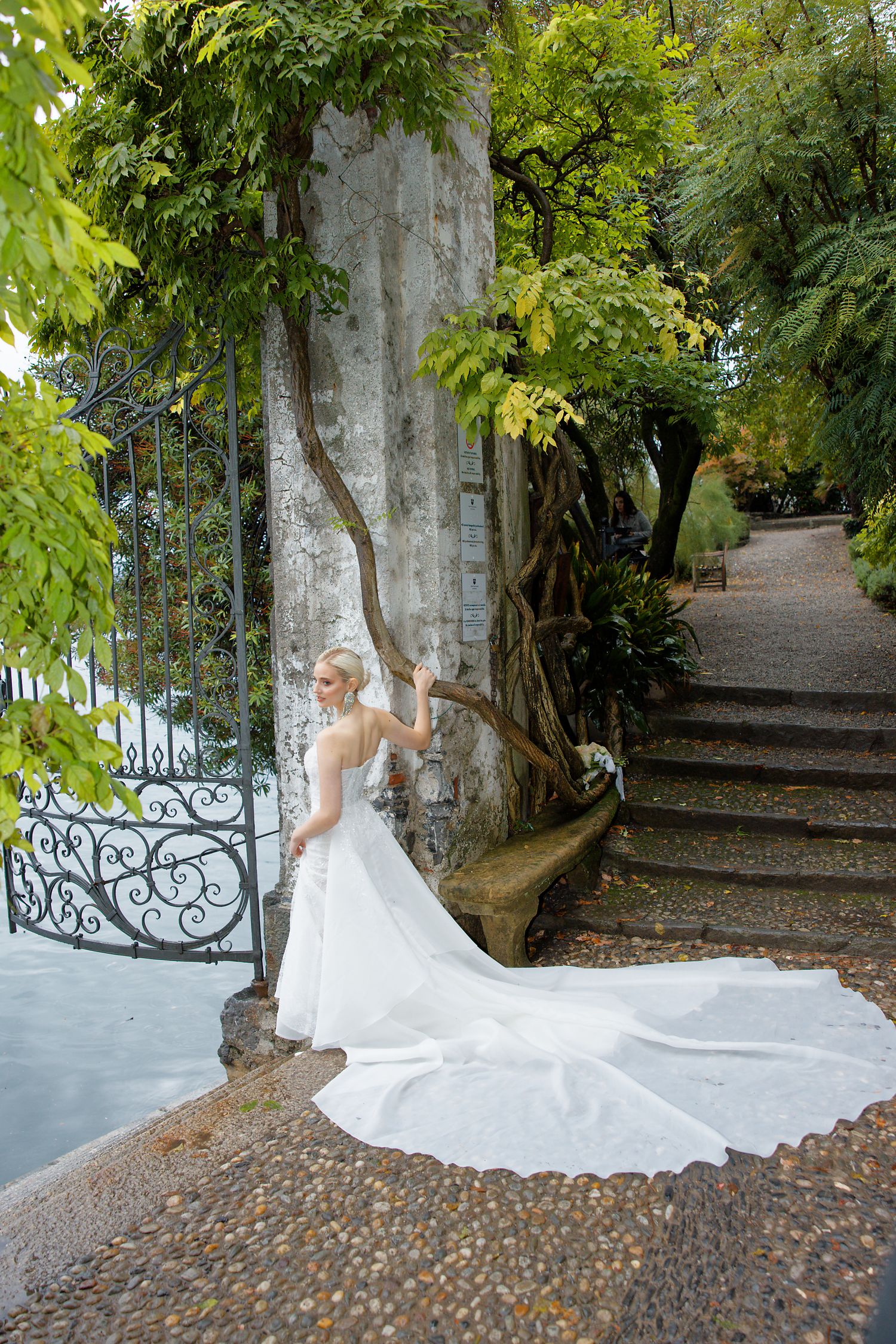 A stunning white wedding dress with a long flowing train spreads across a stone path in a romantic garden setting.