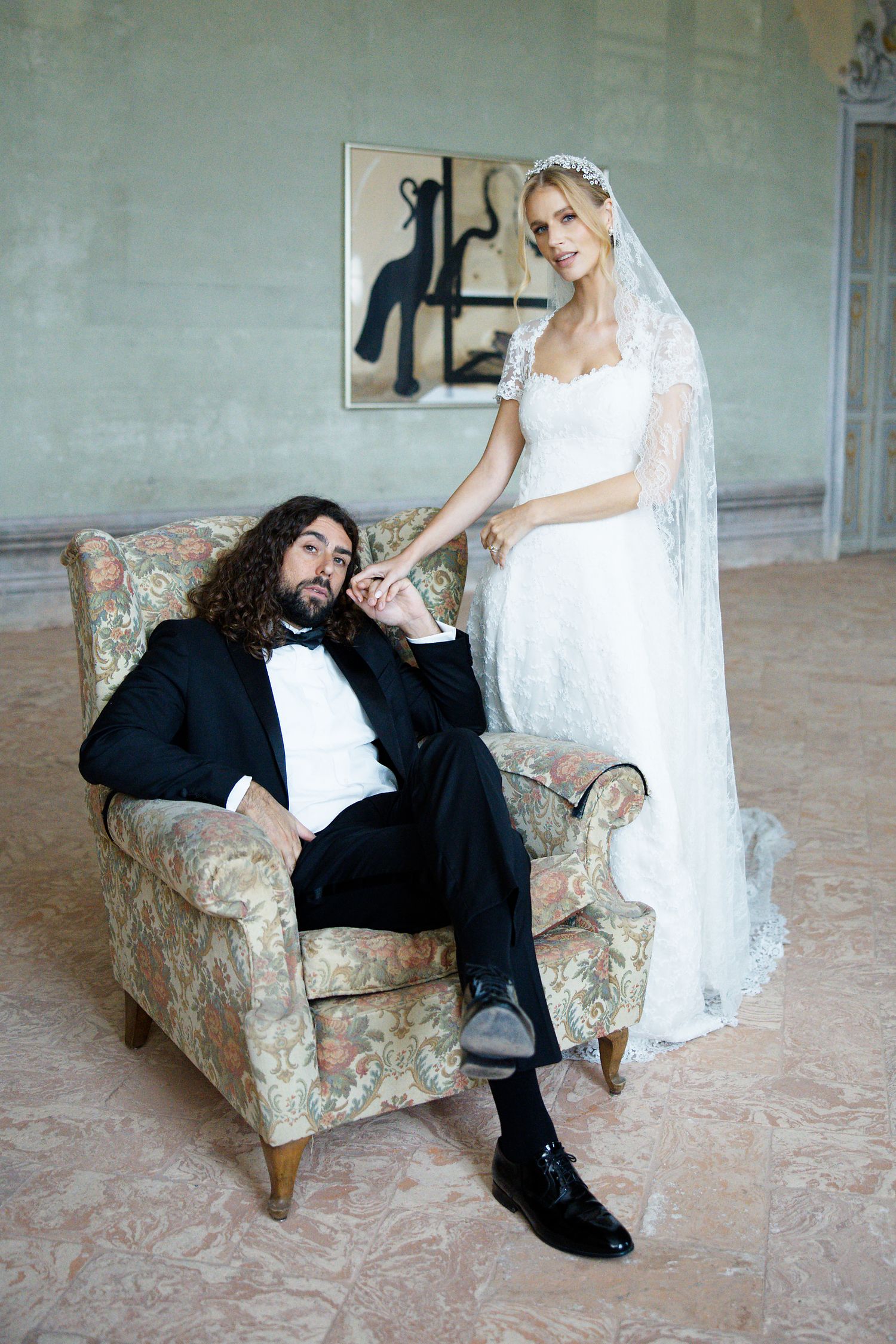 A formal wedding photo showing a person in a tuxedo seated in an ornate chair while a bride in white lace stands nearby.