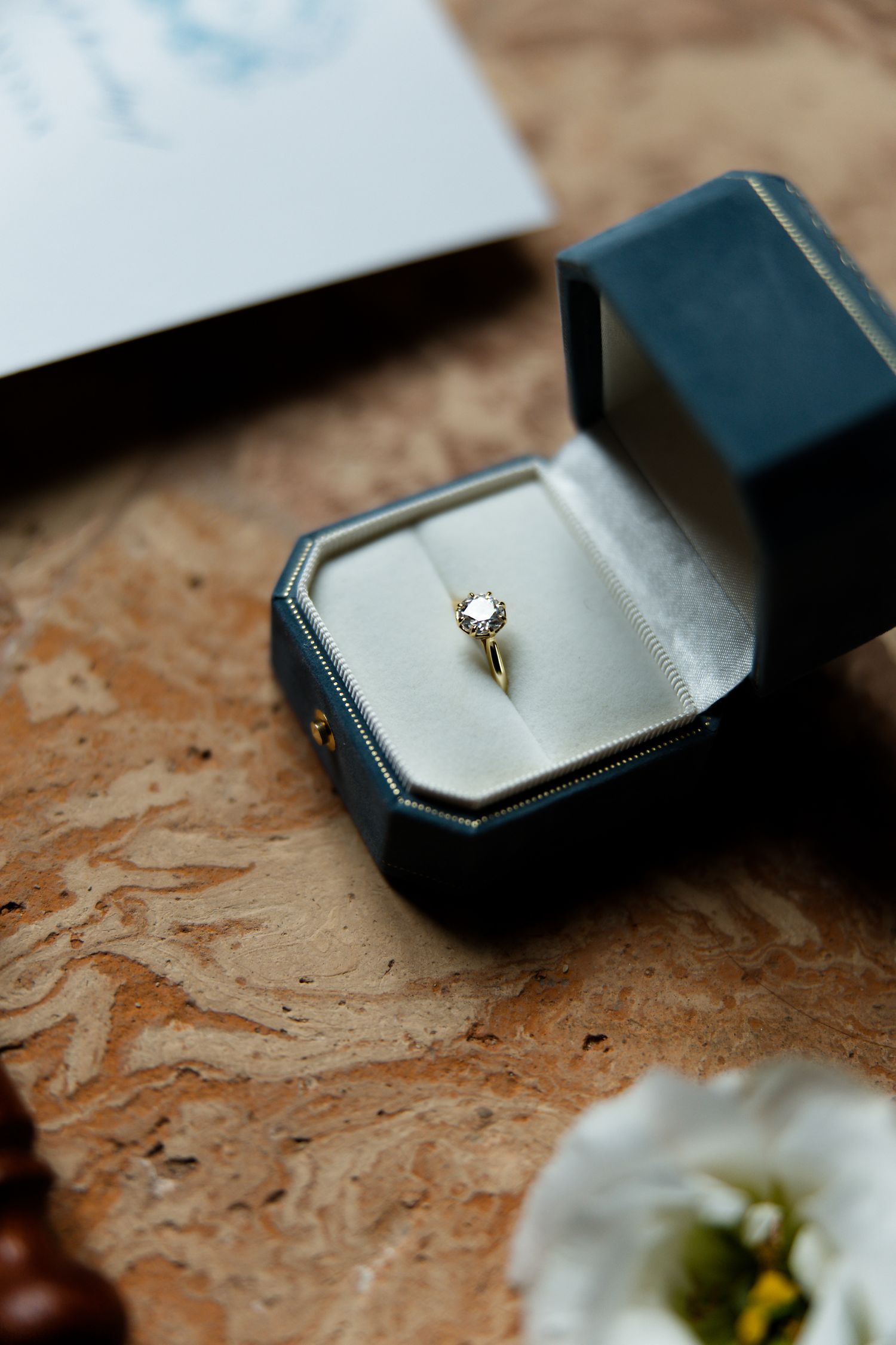 A delicate engagement ring with a solitaire diamond sits in a navy blue hexagonal ring box.