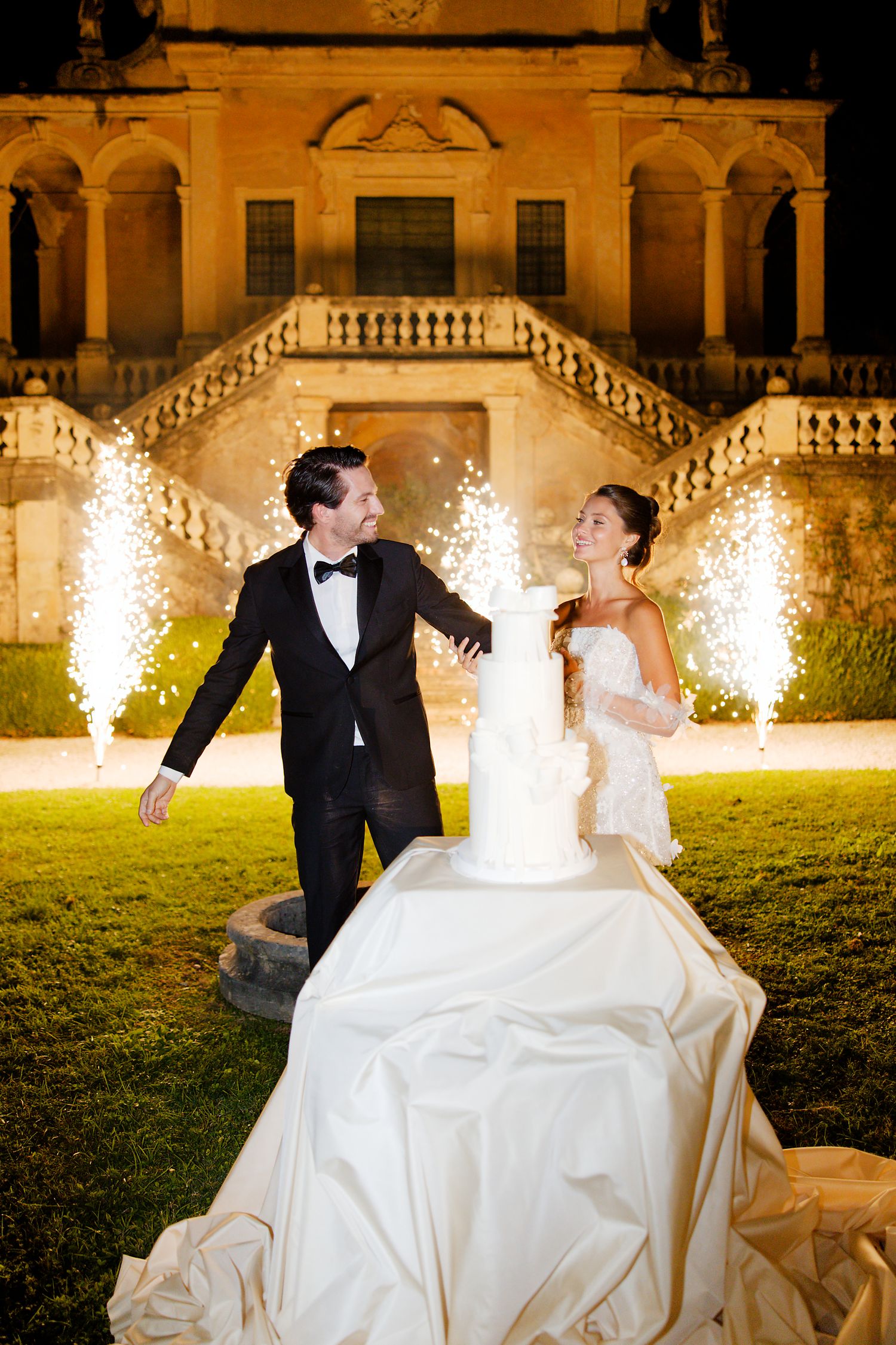 Couple celebrates with sparklers and a towering wedding cake in front of a historic villa.
