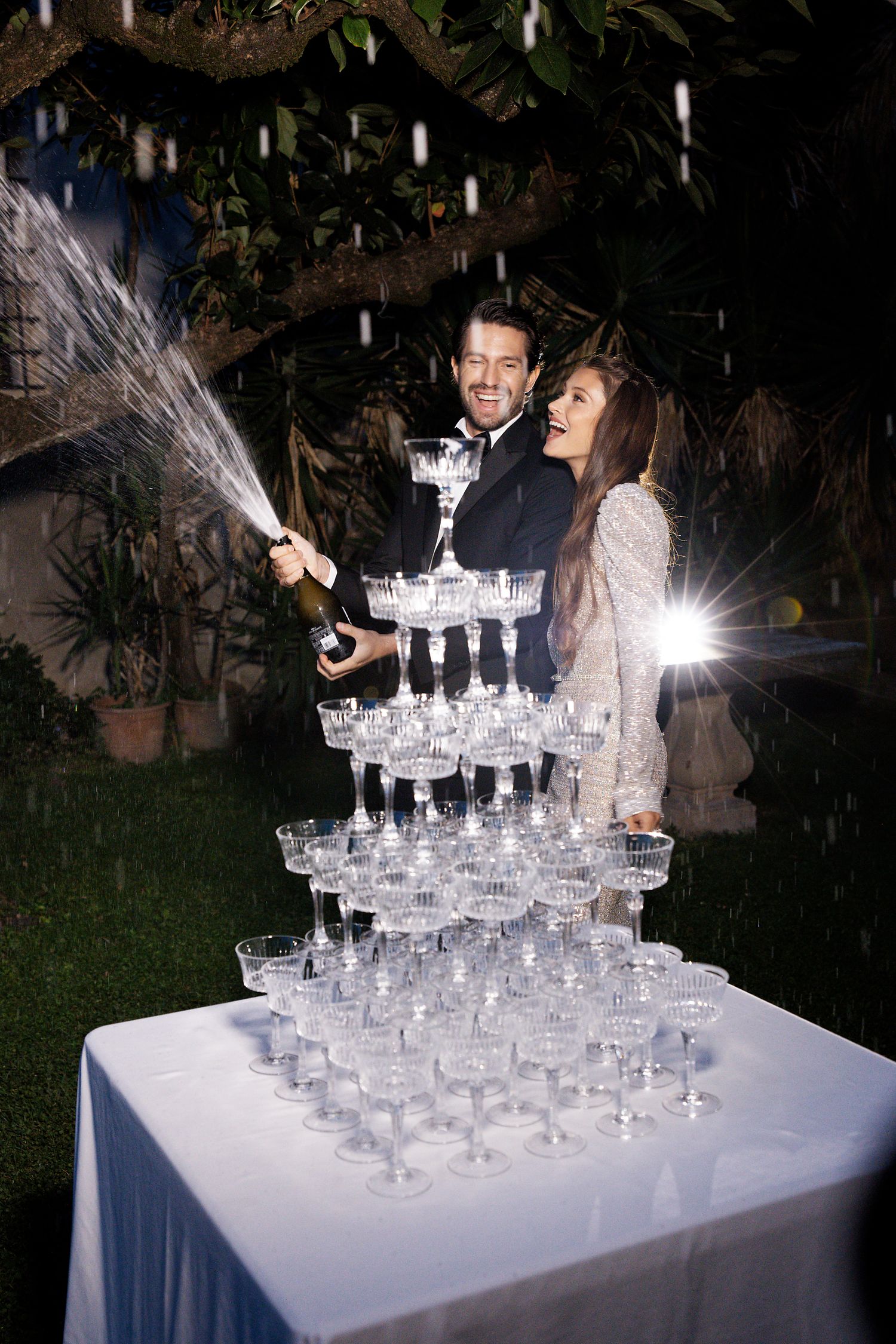 Couple pouring champagne into elegant tower of glasses during nighttime celebration.