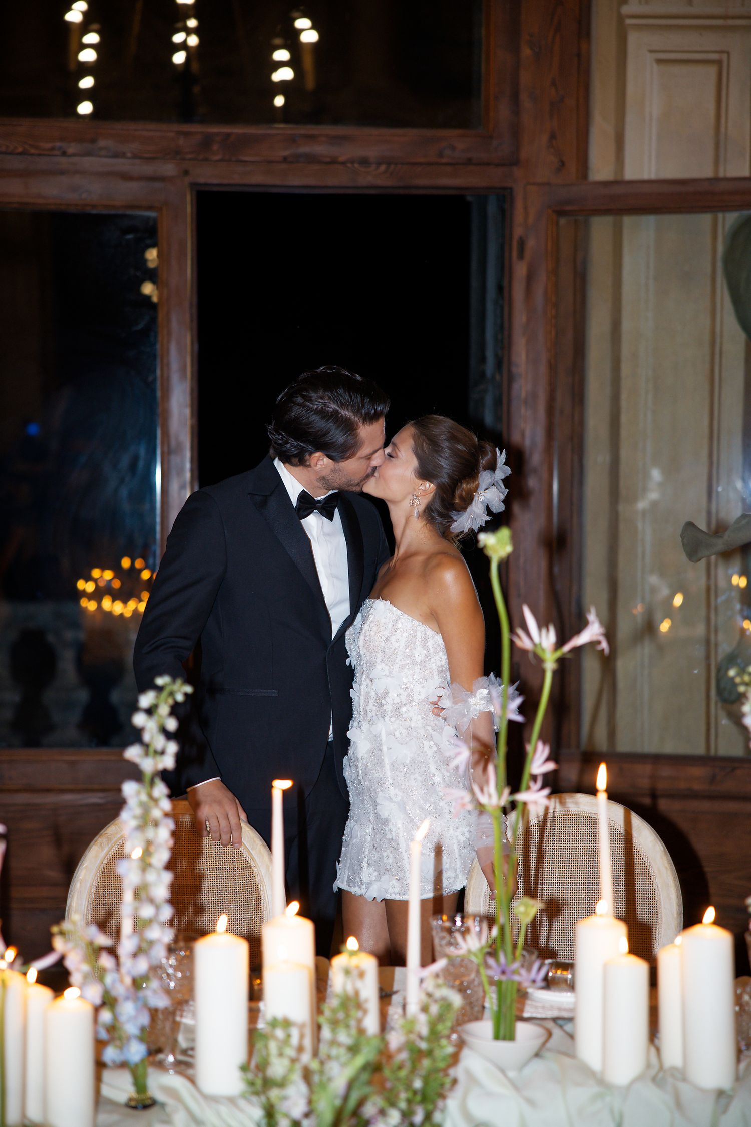 Romantic wedding scene with couple kissing by candlelit table decorated with white florals and glowing lights.