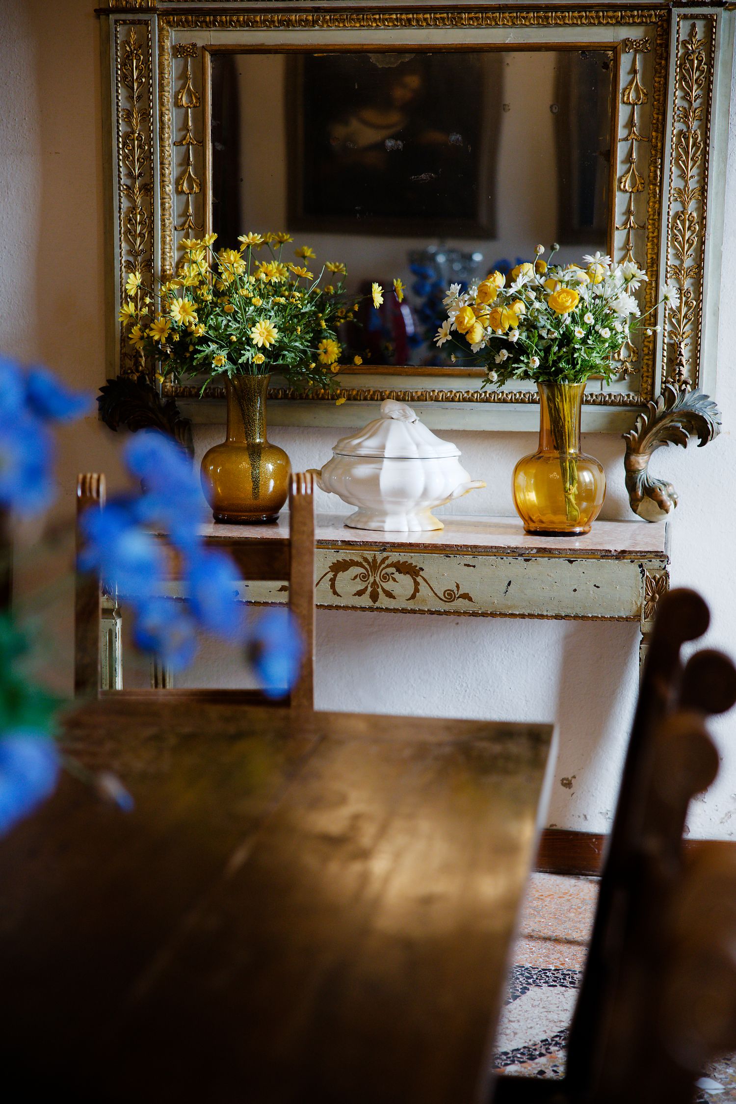 Yellow and blue flower arrangements in amber vases sit atop an ornate console table with a gilded mirror.