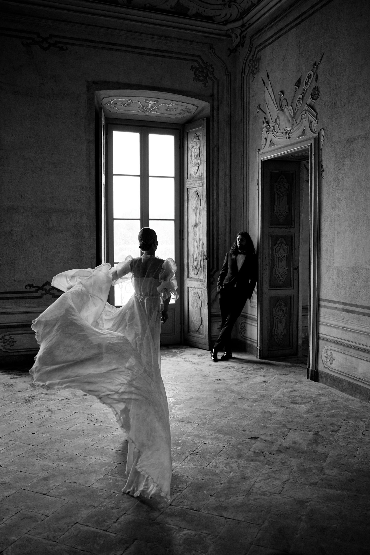 Black and white photo of bride in flowing dress dancing in ornate room while figure stands in doorway.