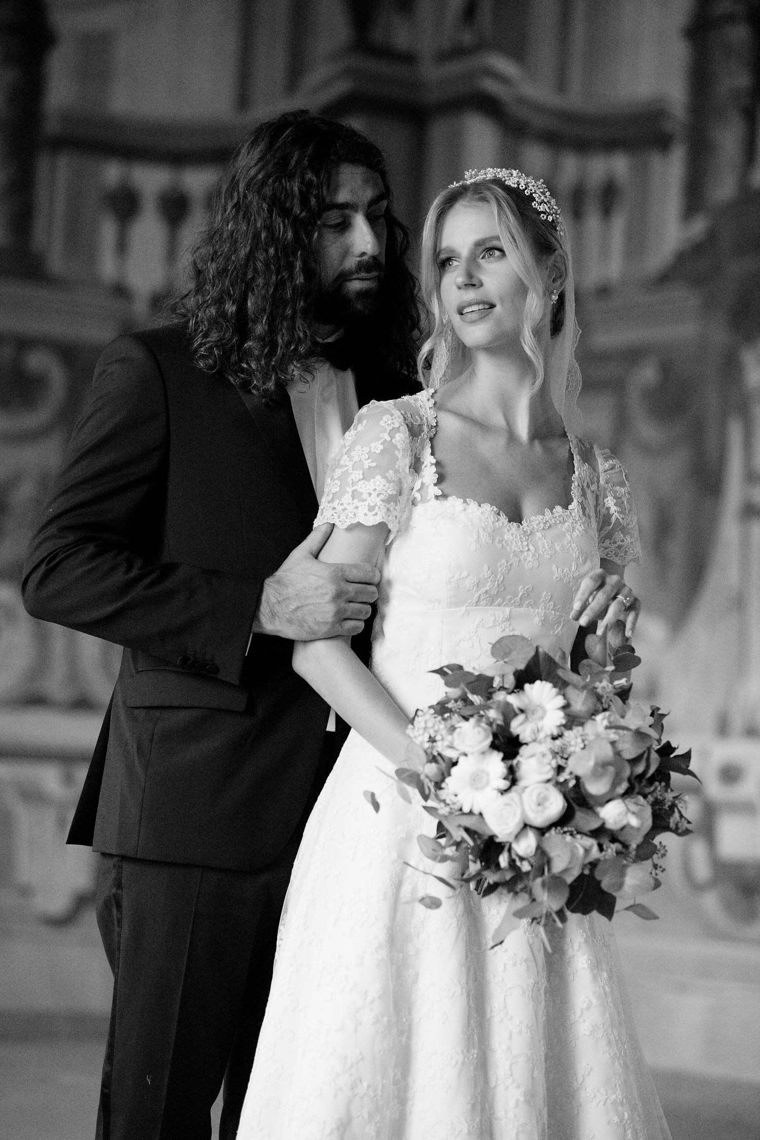 A romantic black and white wedding portrait showing a couple with a beautiful floral bouquet in an elegant setting.