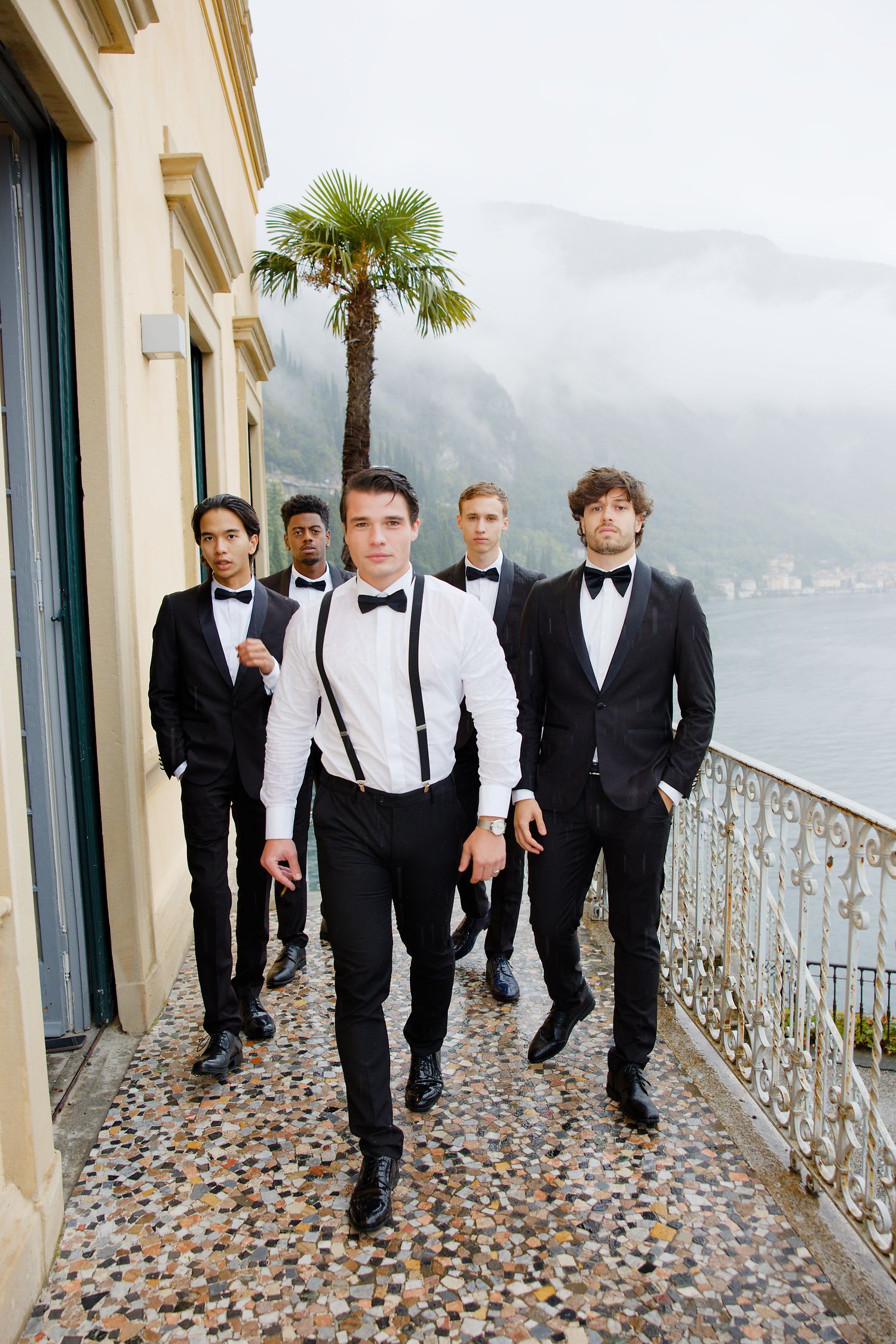 Groomsmen in black tuxedos walking on lakeside terrace with mountain views.