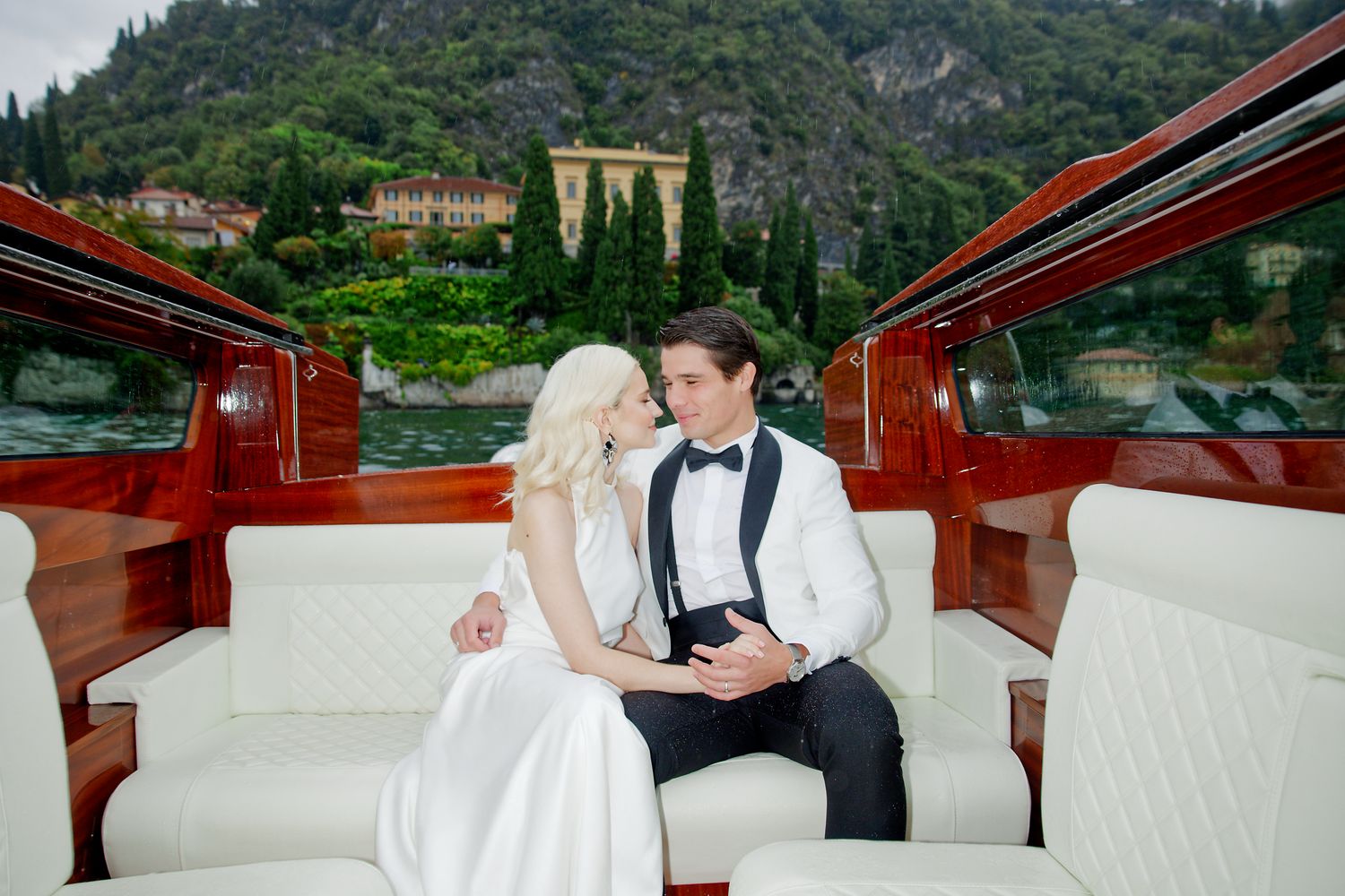 Newlyweds in formal attire sitting close together on classic wooden boat.