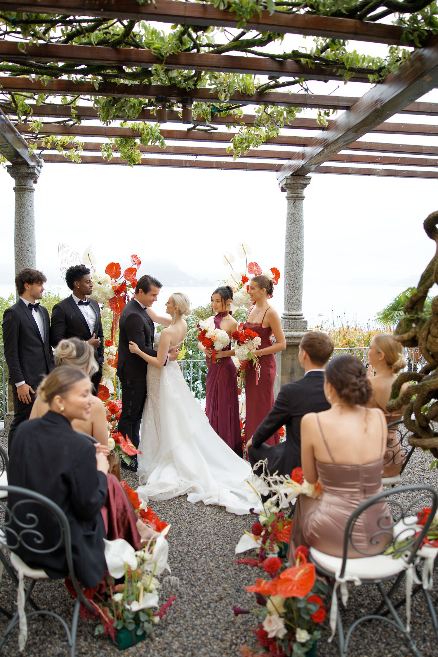 Outdoor wedding ceremony under pergola with orange floral accents and lake view.