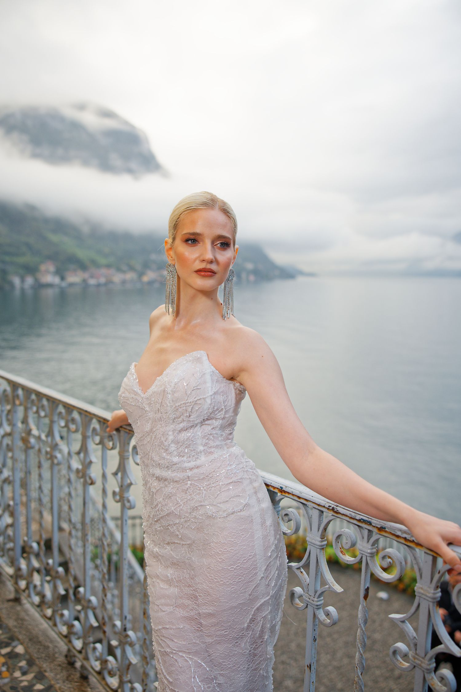 Elegant bride poses against an ornate railing with a scenic lake and mountain backdrop.