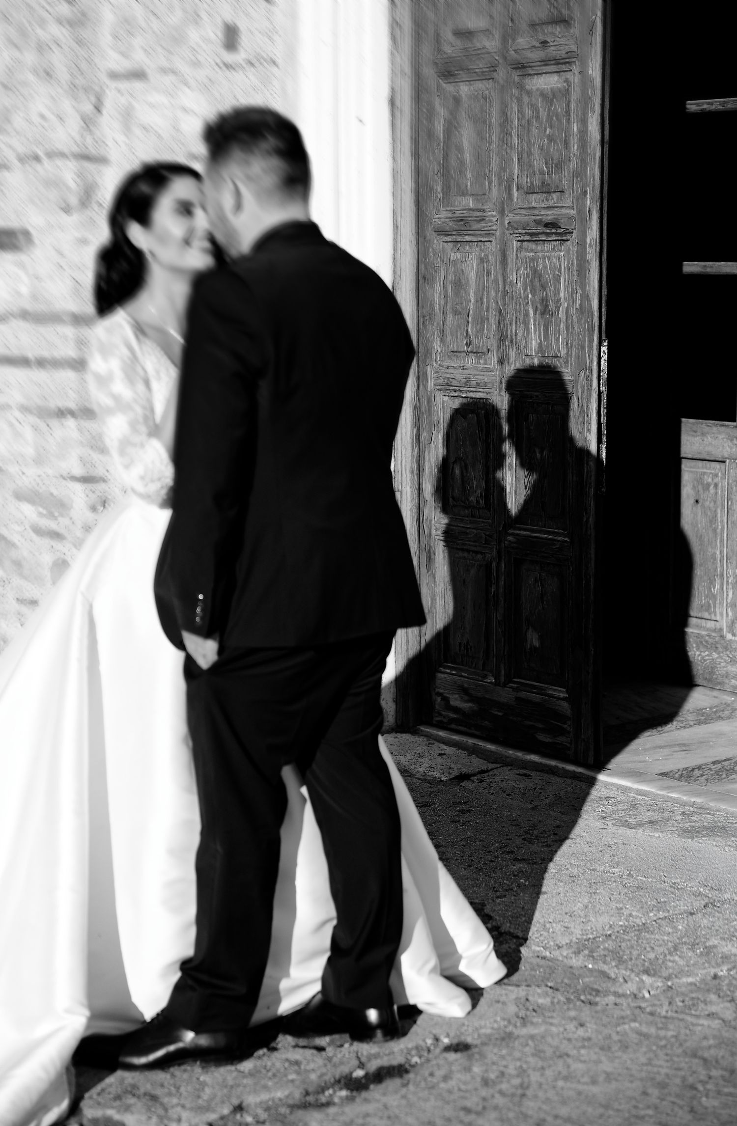 Black and white photo of a bride and groom standing in doorway casting romantic shadows on stone wall.