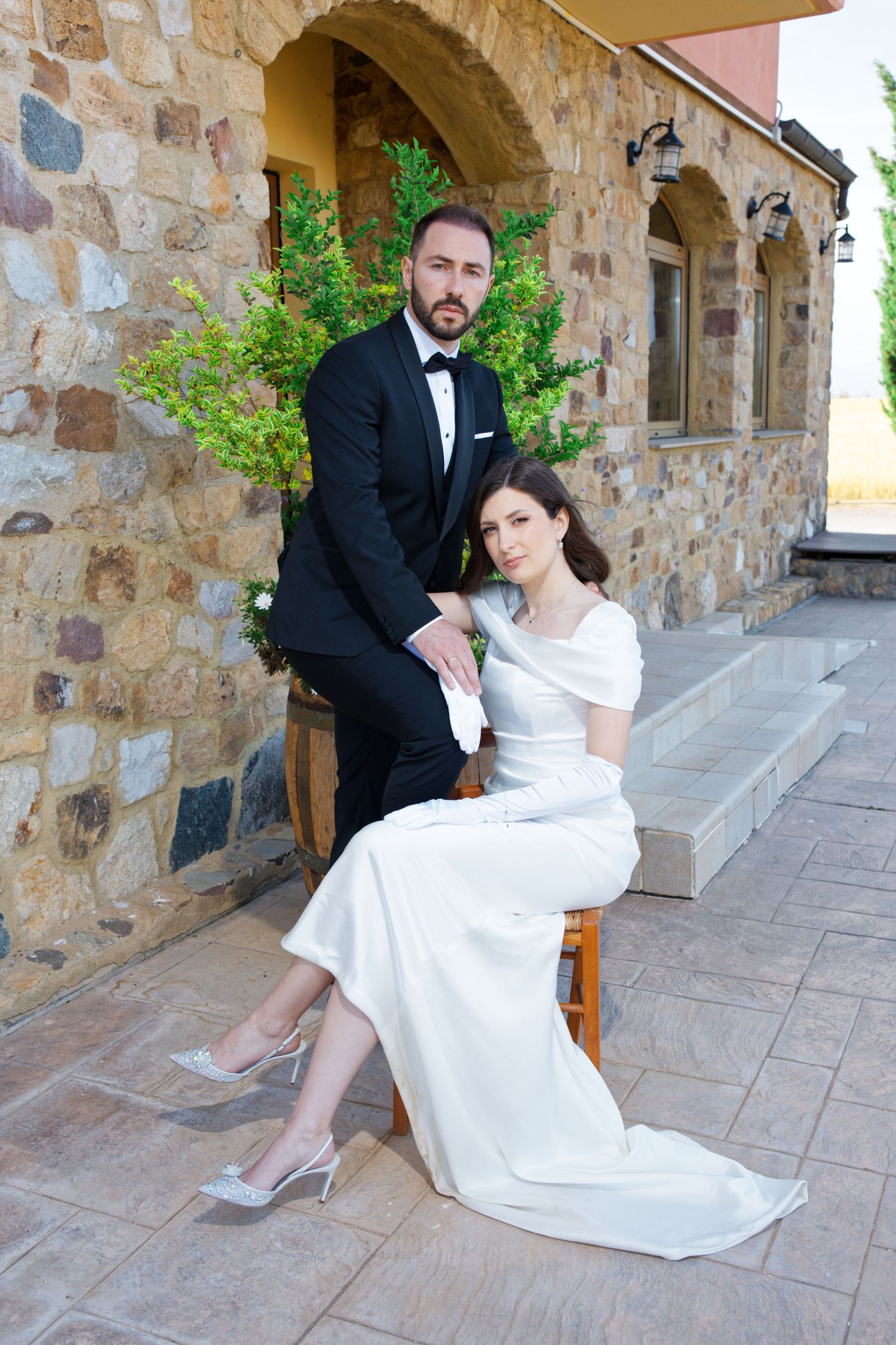 The couple poses elegantly on stone steps outside a rustic Mediterranean-style building.