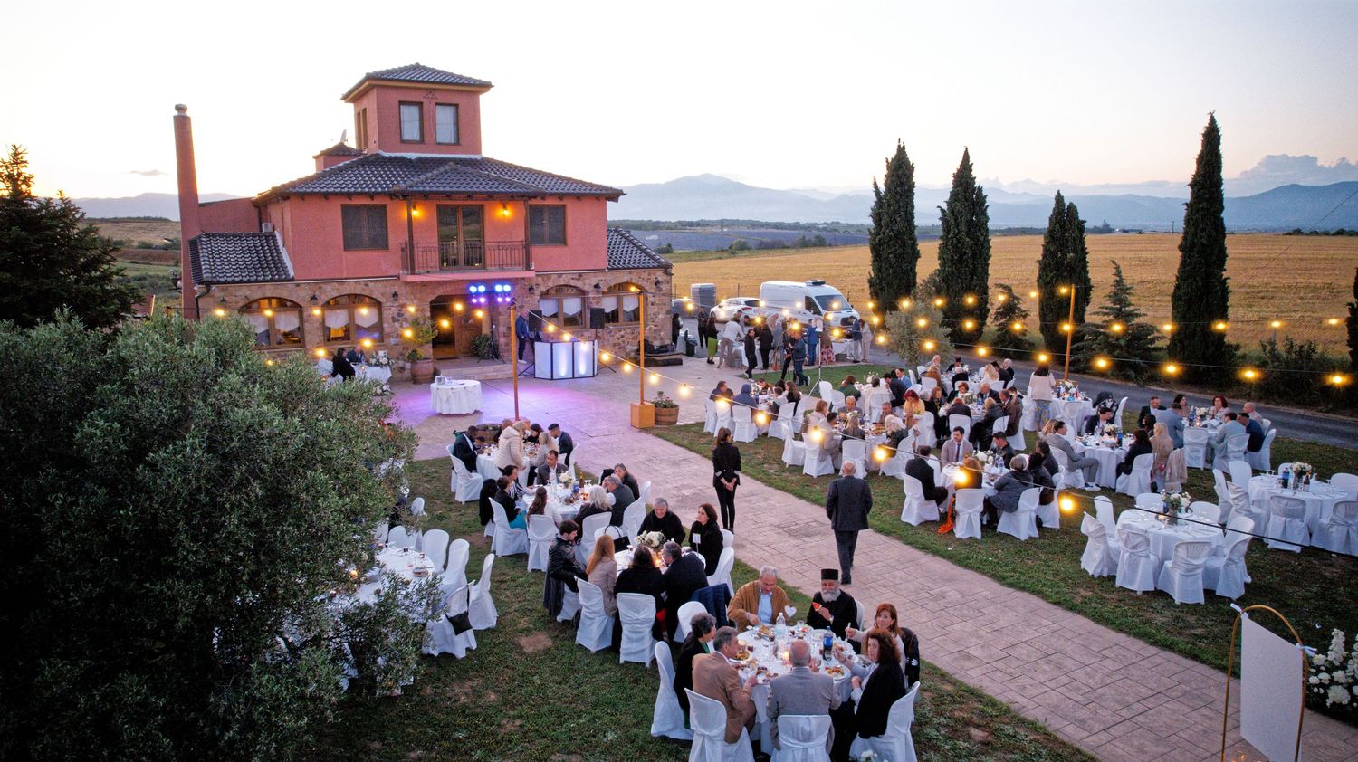 Evening wedding reception at a Mediterranean villa with tables arranged along a lit pathway surrounded by cypress trees.