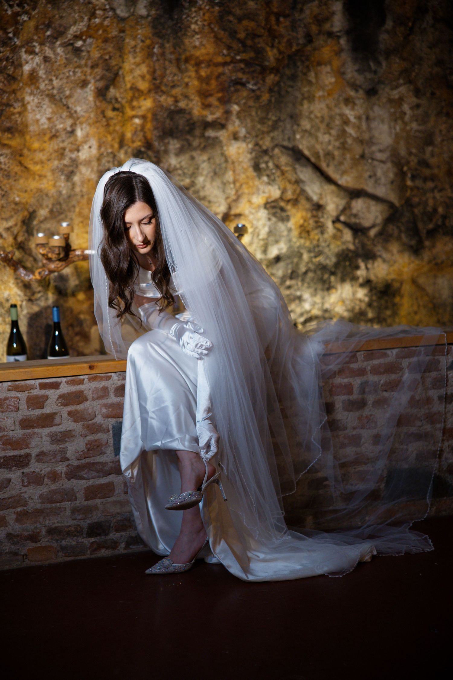 A bride in a flowing white dress and veil poses dramatically against a rustic stone wall.