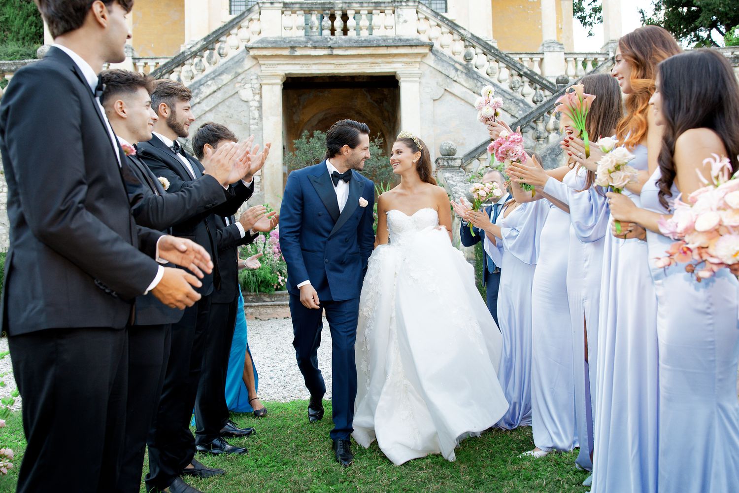 Wedding party celebrates on lawn with bridesmaids in blue dresses and groomsmen in black tuxedos.