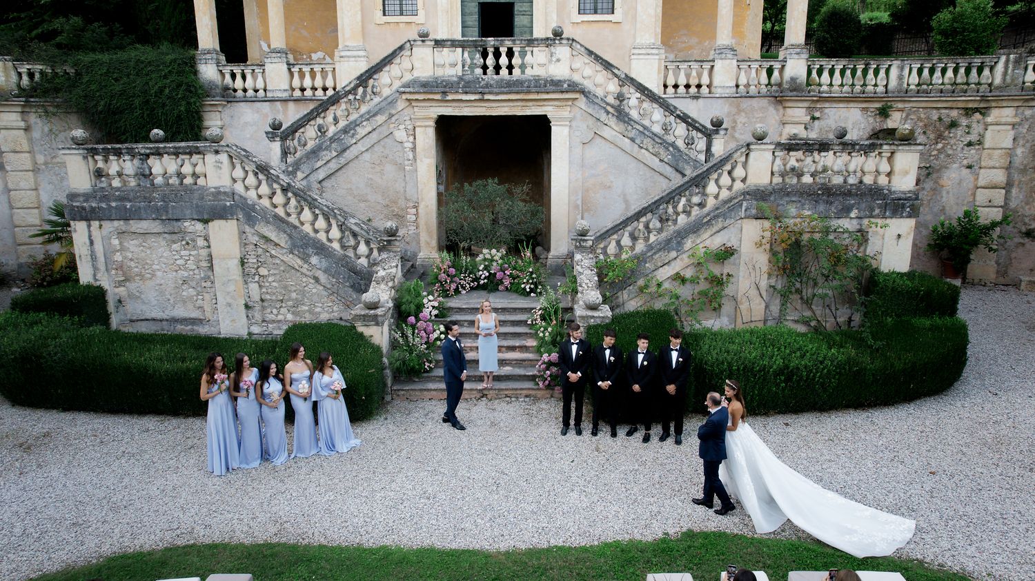 Wedding party in formal attire stands on gravel path in front of elegant stone staircase at Italian villa.