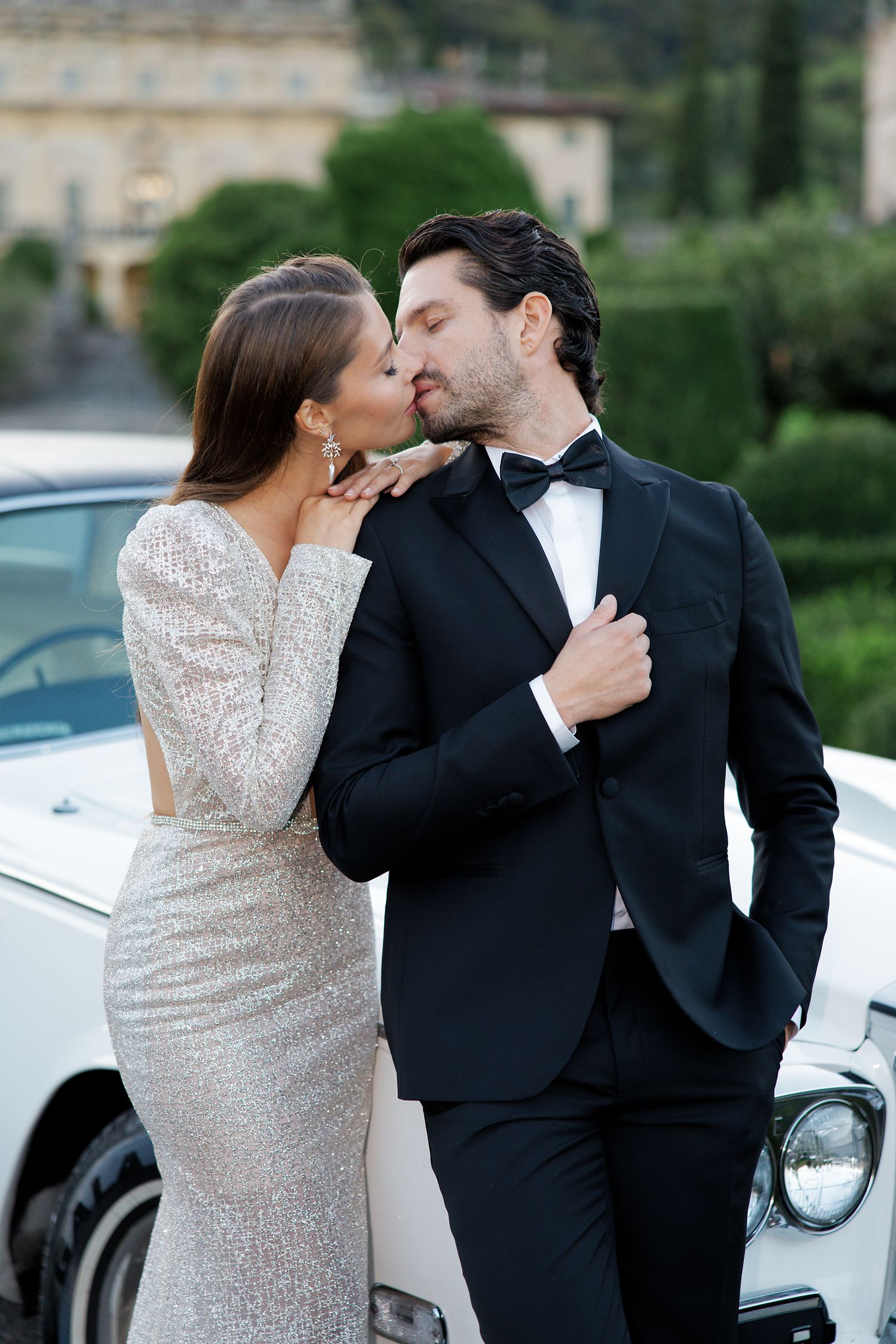 A romantic couple in formal attire share a kiss while posing next to a classic white car.