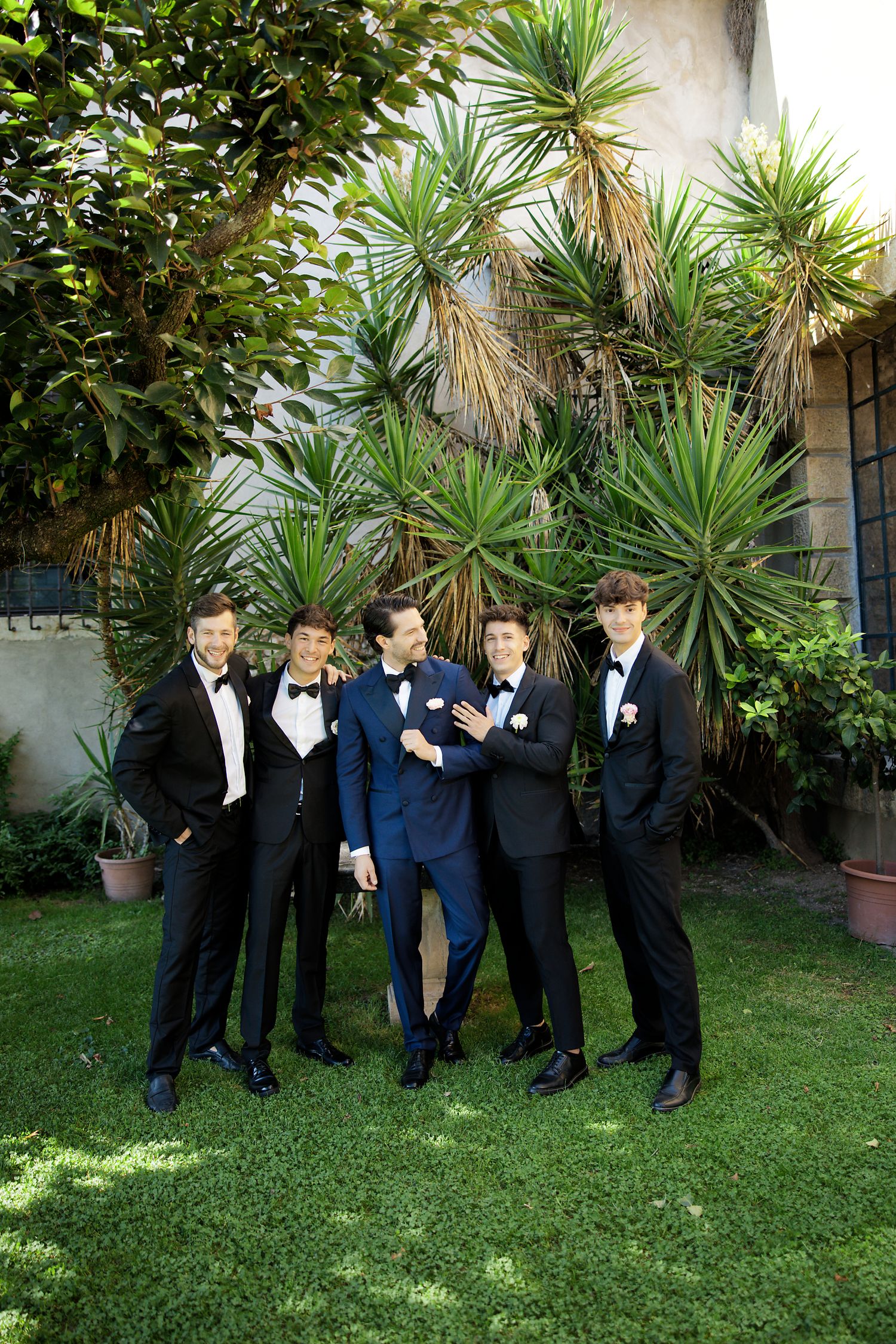 Groomsmen in formal attire pose together in front of tropical plants and palm trees in an outdoor garden setting.