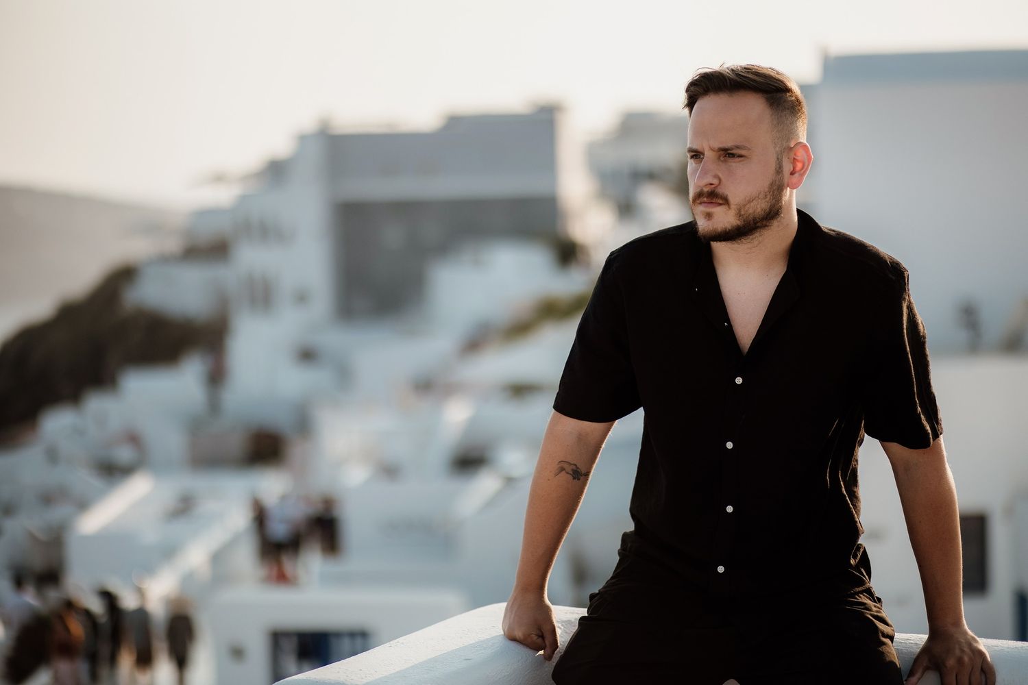 A person in a black button-down shirt stands against a backdrop of white buildings during golden hour.