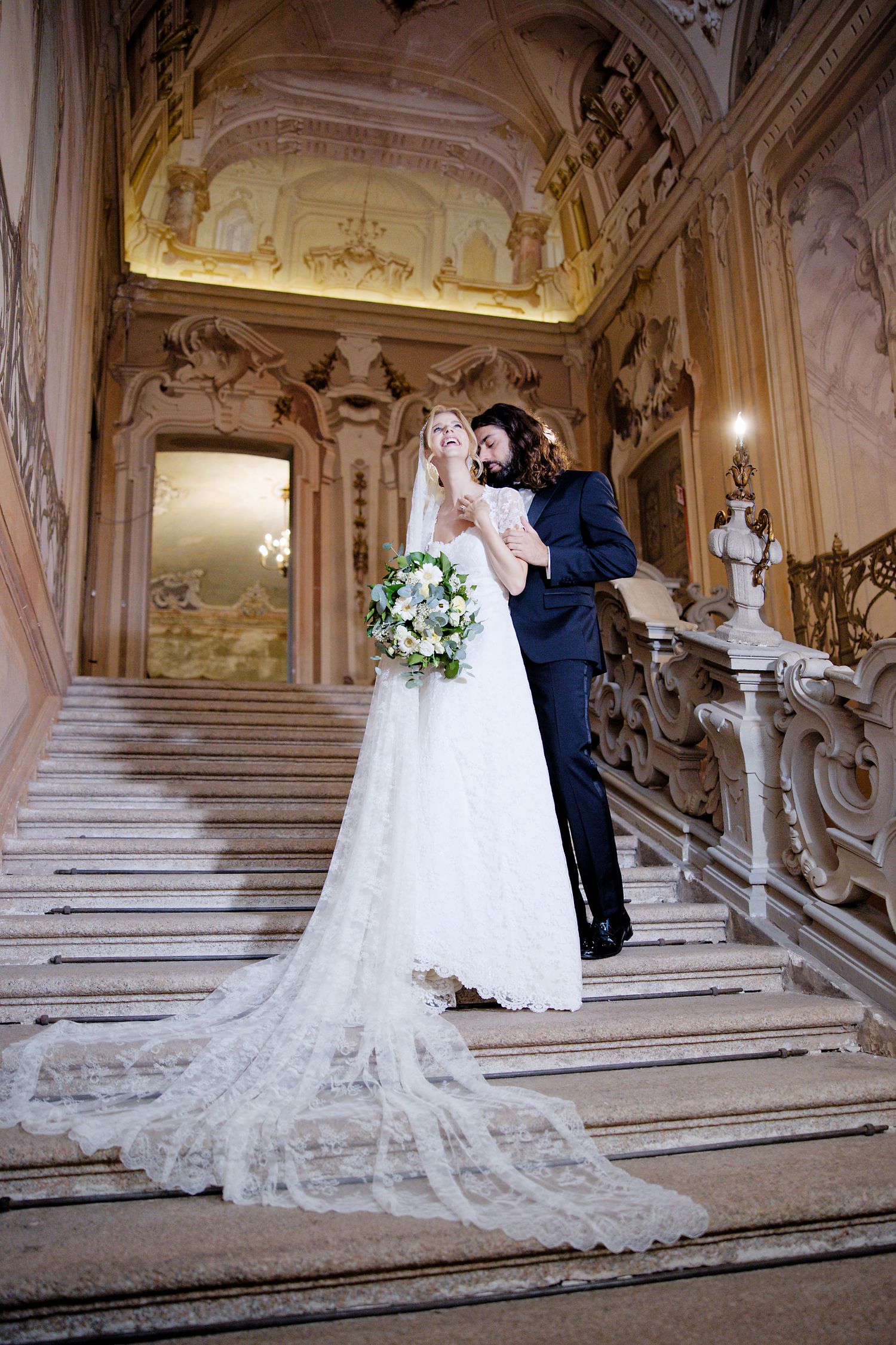 Elegant wedding couple poses on ornate marble staircase in historic palace interior.