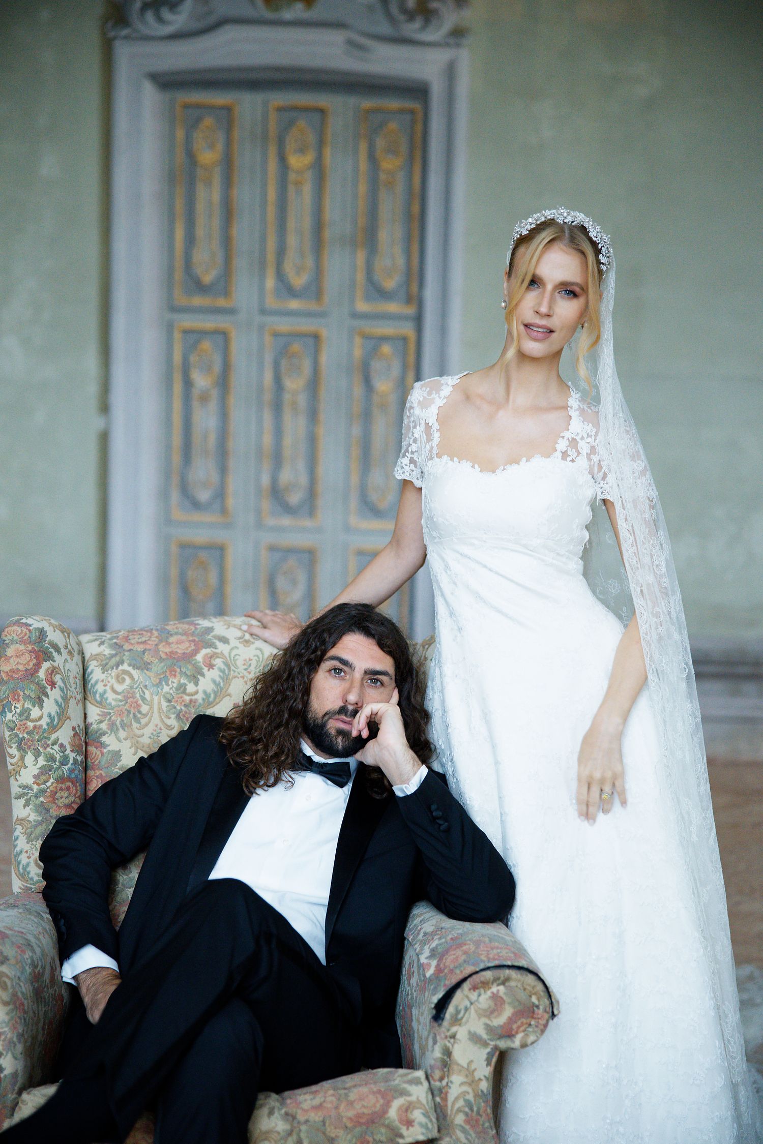 A wedding portrait featuring a bride in white lace dress and groom in tuxedo lounging on a vintage floral armchair.
