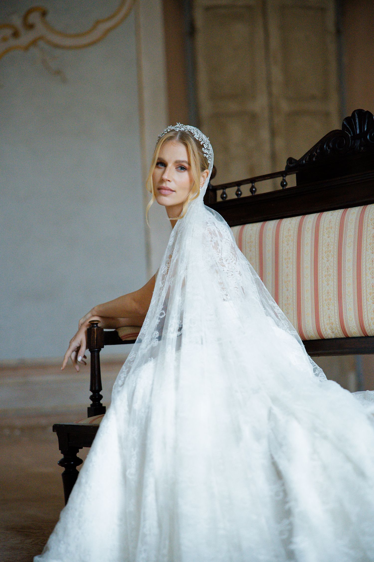 A bride wearing a delicate lace dress and headpiece sits on an antique striped bench in an ornate room.