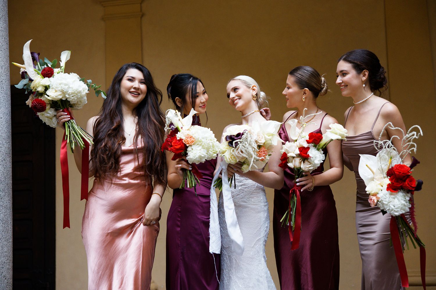 A wedding party in elegant gowns poses together holding bouquets of white and red flowers.