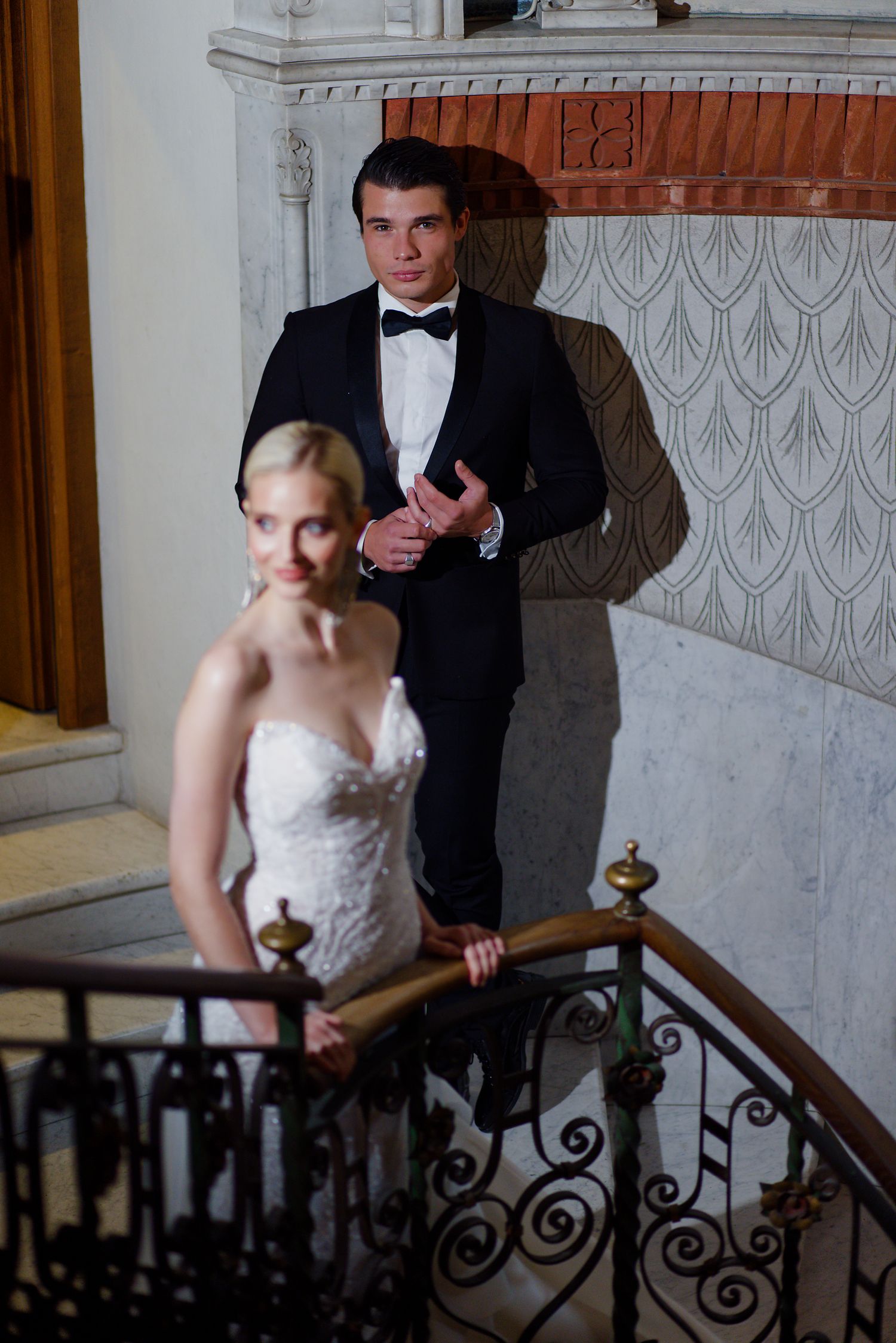 A couple in wedding attire poses on an elegant staircase with ornate wallpaper details.