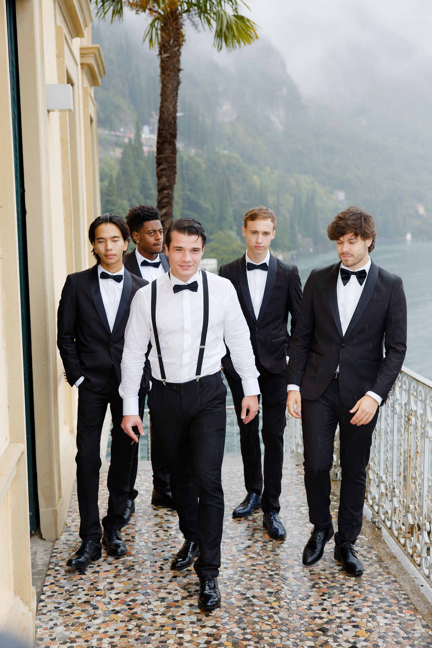Five groomsmen in black tie attire walk along a scenic balcony with mountain views.