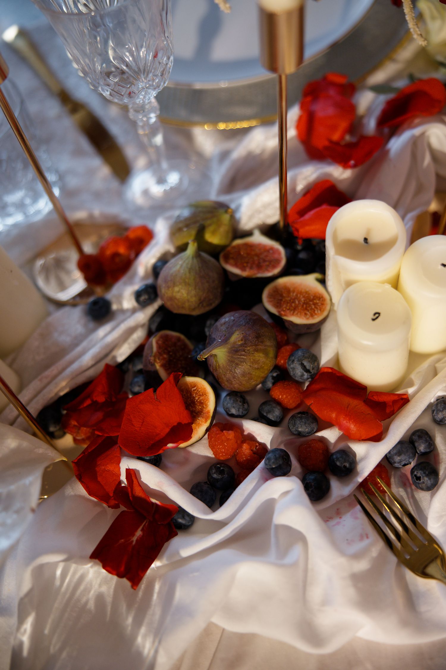Elegant table setting with fresh figs, blueberries, and white candles on silk tablecloth.